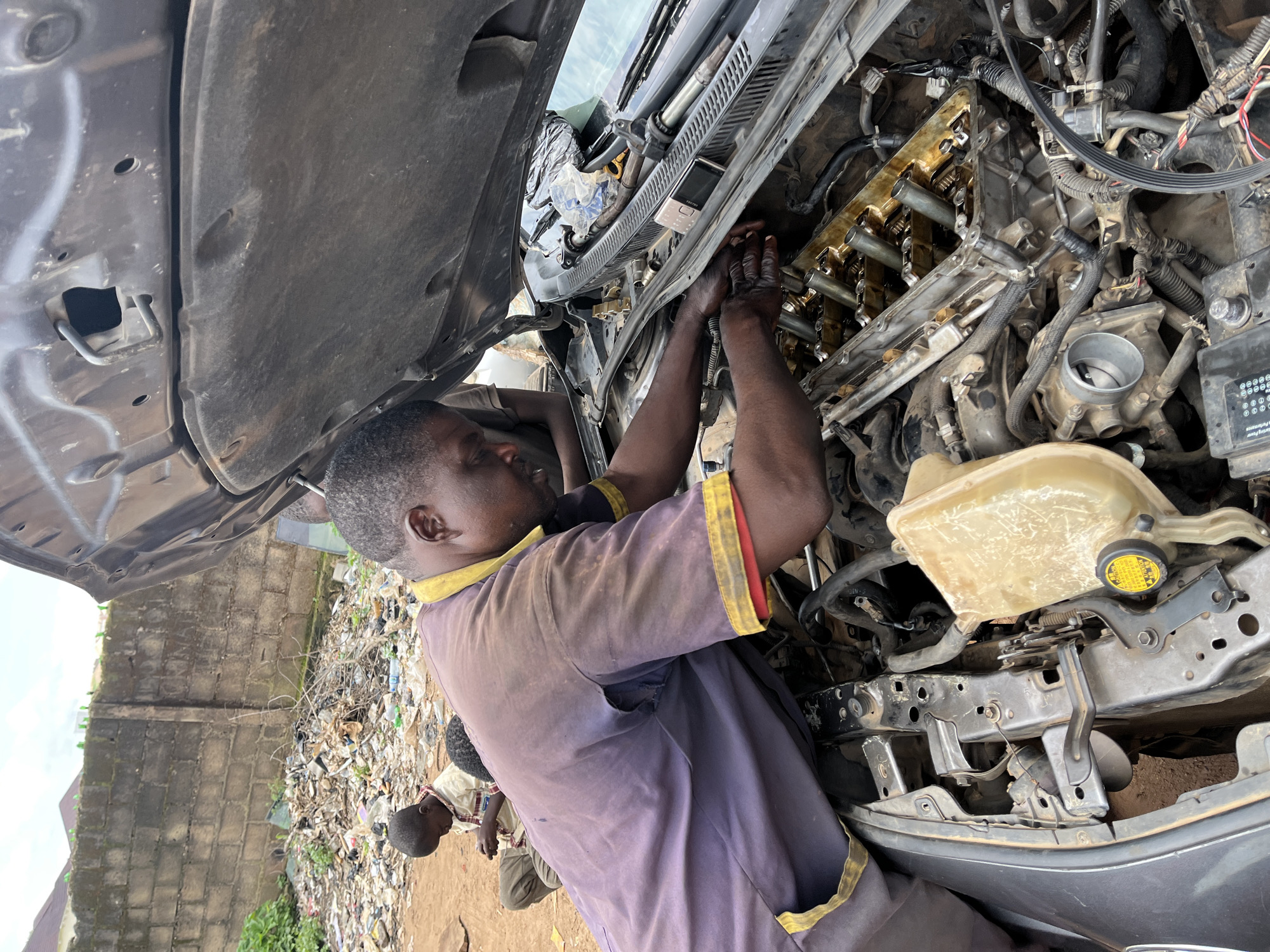 An outdoor scene in Jos, Nigeria, shows a mechanic working on a car engine. The mechanic, a Black man wearing a purple and yellow mechanic's shirt, is hunched over the open hood of a dark-colored vehicle, his hands manipulating parts of the engine. The engine is partially disassembled, with various components and wiring exposed. To the left of the mechanic, a young boy in a checkered shirt and brown pants sits on the ground, looking down at something in his hands, possibly a mobile phone. Behind them, a pile of debris and trash is visible next to a rough, textured brick wall. The ground appears to be dirt. The sky above is overcast.