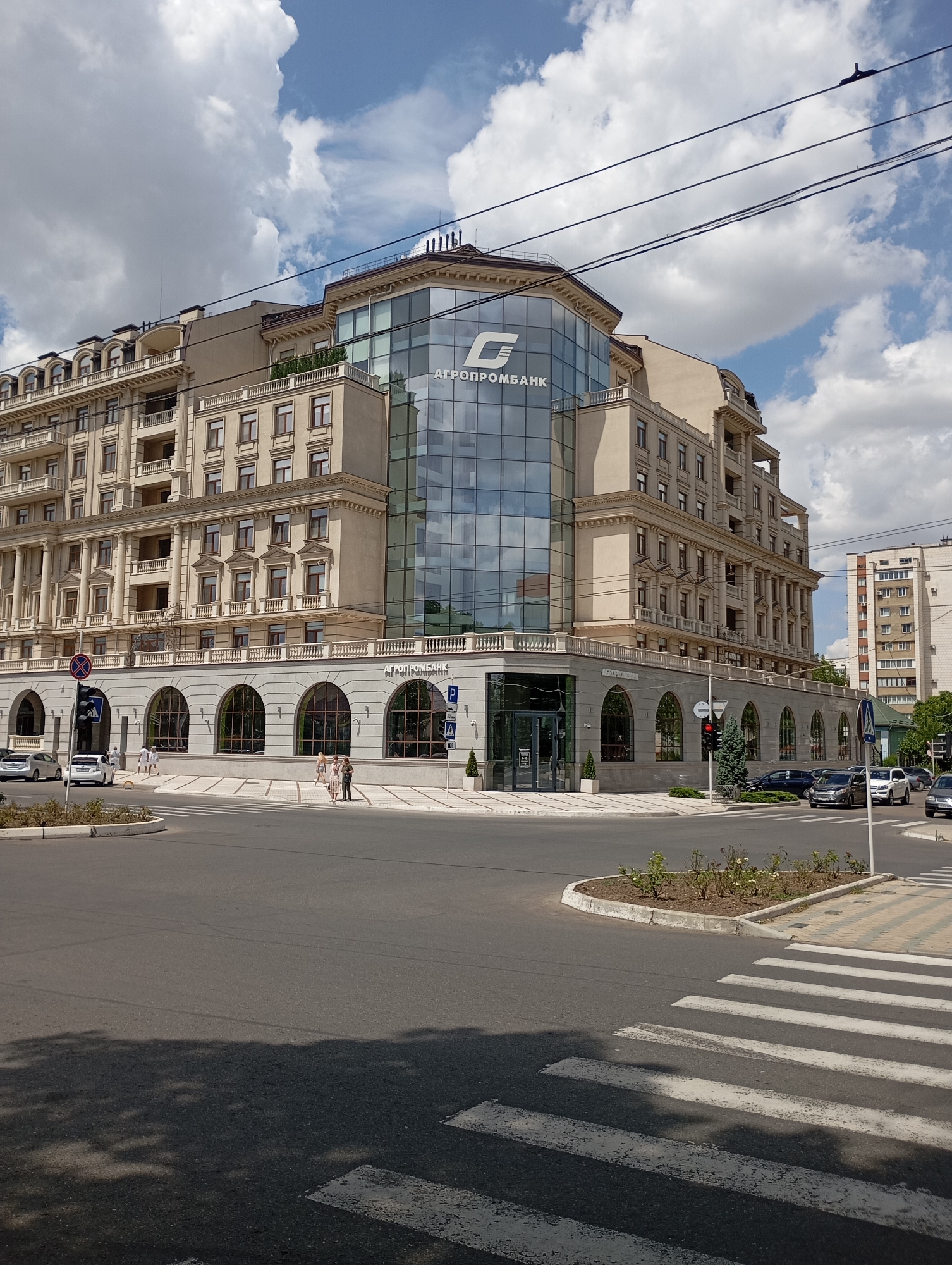 The image captures a street view in Tiraspol, Moldova, focusing on a large, multi-story building with a modern glass facade on the upper floors. The building features a combination of classical and contemporary architectural styles, with ornate detailing on the lower, beige-colored sections and a sleek, reflective glass structure on the upper corner. A prominent logo, possibly for a bank named "AgroPromBank," is visible on the glass section.
The scene is set at an intersection with a pedestrian crossing in the foreground. Several cars are present on the street, including a white sedan and a dark-colored hatchback on the right side of the intersection, and a white sedan and a silver hatchback on the left. A traffic light and street signs are visible, indicating an urban environment.
A few pedestrians are scattered around the scene. Two individuals are standing near a building entrance on the right side of the image, while a small group of three people are walking on the sidewalk in front of the building's arched ground-floor windows. Two other individuals are visible further down the sidewalk on the left.
The sky is partly cloudy with large, white cumulus clouds against a blue backdrop. Power lines are strung across the upper portion of the image. The overall impression is of a bright, daytime scene in a city. A taller, older residential building with multiple balconies and windows can be seen in the background on the right. Trees and some landscaping are present around the building and at the edge of the intersection.