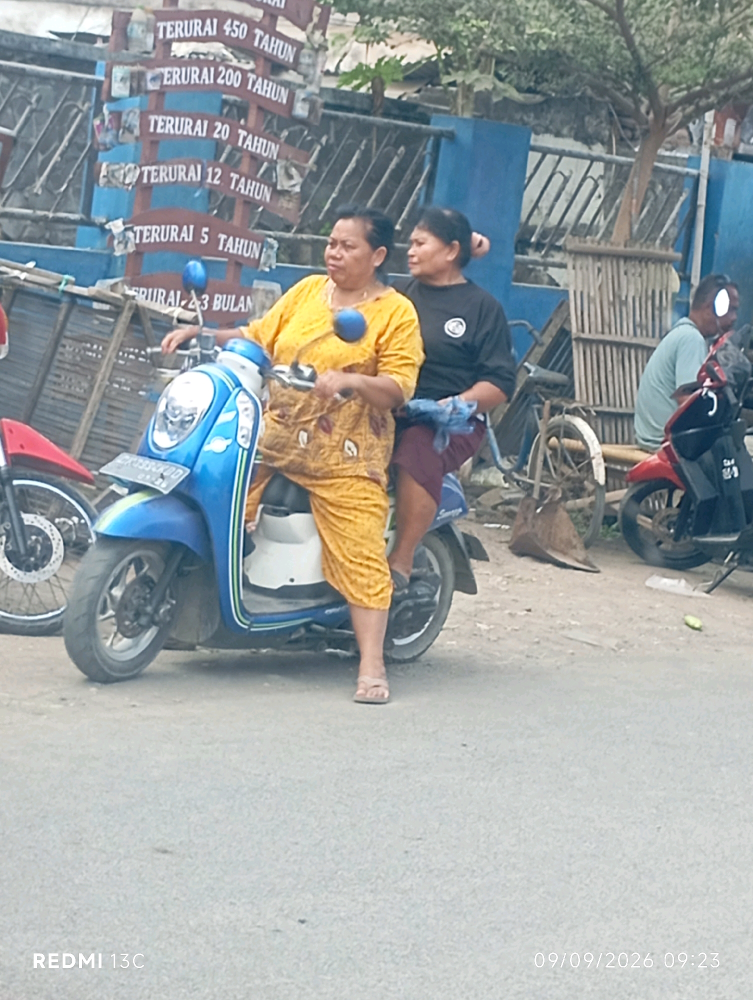 The image depicts a street scene in Wuluhan, Indonesia, with a focus on two women on a blue and white scooter. The woman in front, wearing a yellow patterned outfit, is driving. Behind her, a woman in a black shirt is seated. They are on a paved surface, likely a road, with other vehicles and a blue fence visible in the background. Signs with Indonesian text, indicating durations in years and months, are mounted on a structure to the left of the scooter. To the right, another scooter with a red body is parked. The overall environment suggests an outdoor, possibly commercial or residential, area within a town.
