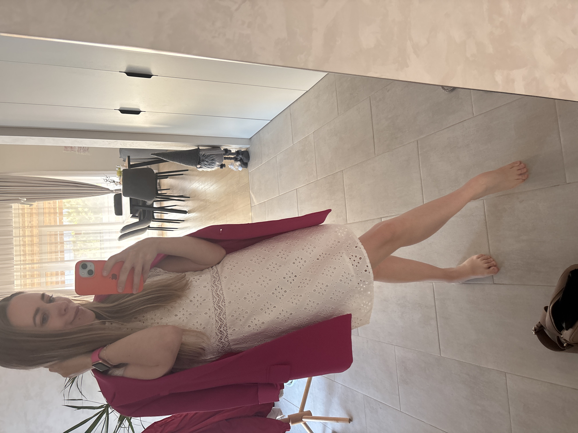 A woman is taking a selfie in front of a mirror. She is wearing a white dress and a bright pink blazer. She is holding an orange iPhone. The location appears to be indoors, possibly in an apartment or house. In the background, there is a cabinet, a dining table with chairs, and tiled flooring. A bag lies on the floor near her bare feet. The specific location in Chaikovsky, Russia cannot be determined from the image.