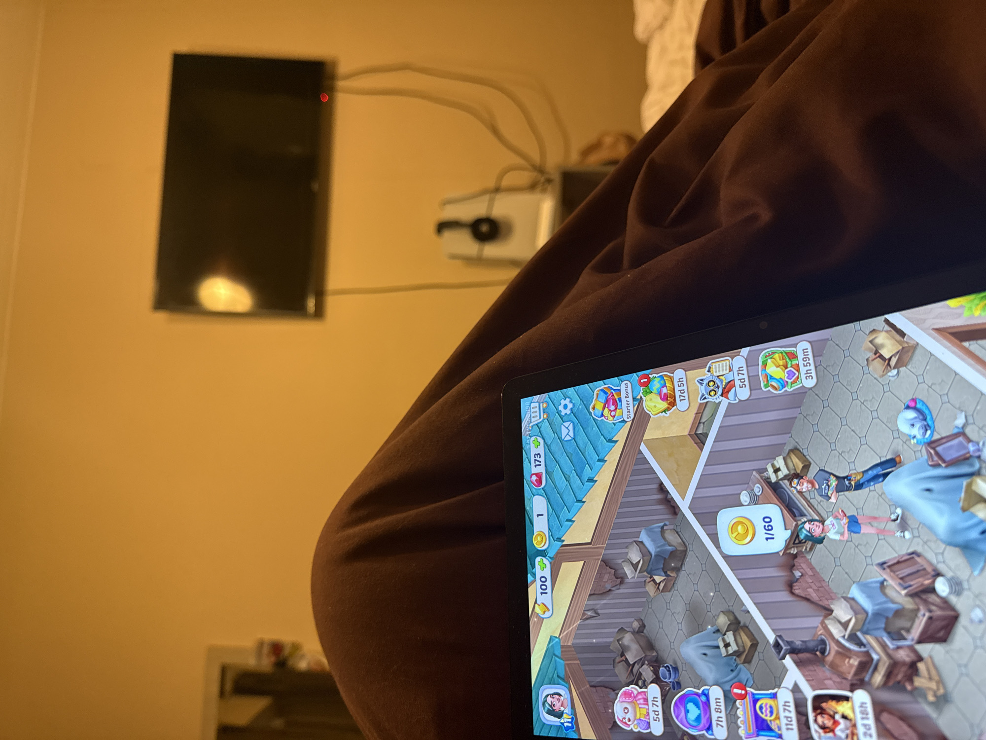 A low-angle shot depicts a person sitting in a soft, brown garment, with a tablet resting on their lap displaying a mobile game. In the background, a flat-screen television is mounted on a yellow wall, accompanied by visible cables and a white device. The room appears to be dimly lit. The game on the tablet features cartoon-style characters and interior design elements.