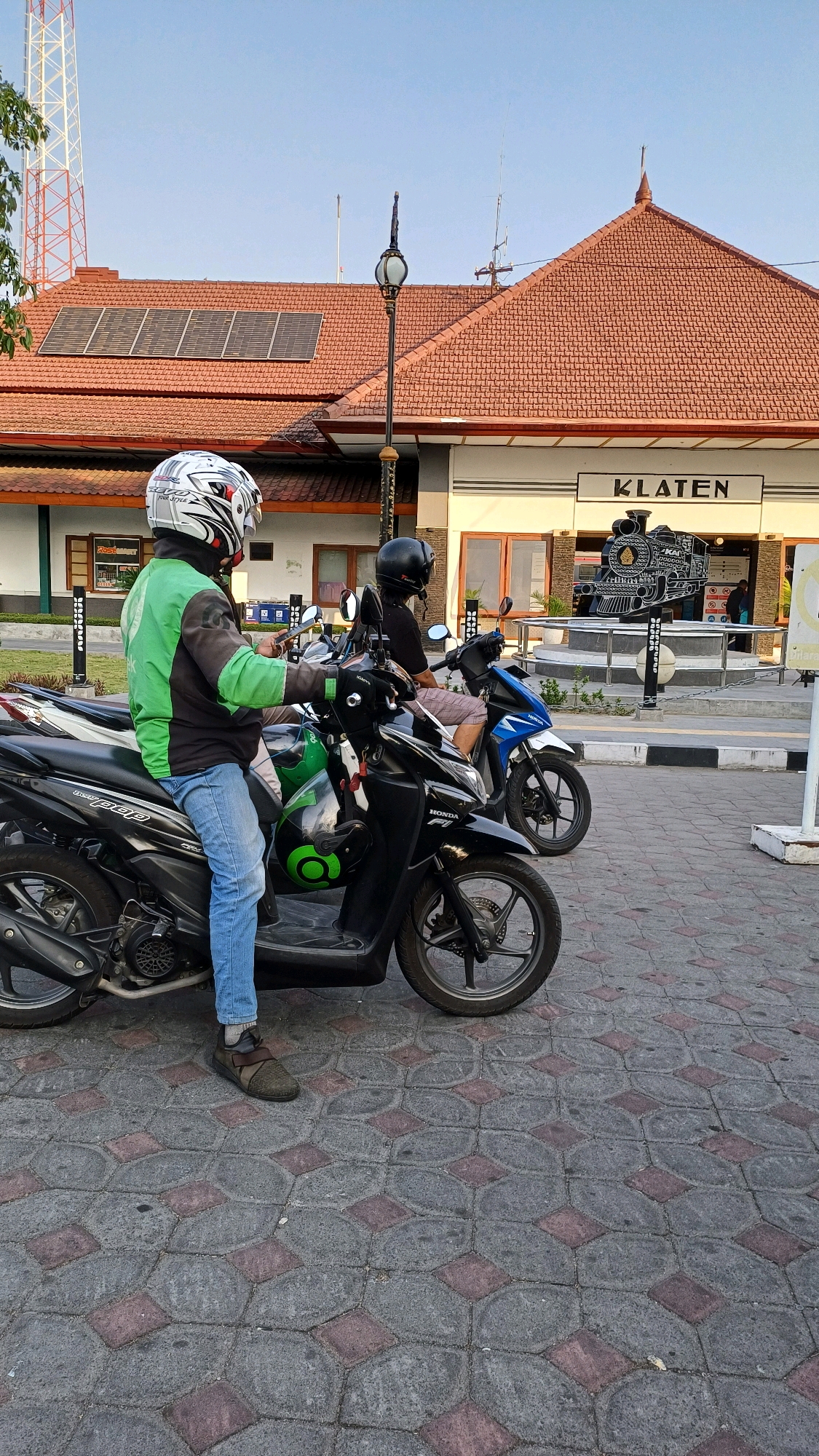 The image depicts an outdoor scene in Klaten, Indonesia, likely near a train station given the prominent sign that reads "KLATEN" above an artistic representation of a steam locomotive. In the foreground, two individuals are seated on motorcycles. The person on the left, facing right and slightly away from the camera, wears a green and black jacket, blue jeans, and a white helmet adorned with red and black graphics. This individual appears to be holding a smartphone. The motorcycle they are on is black and has a green circular logo on its side. To the right of this motorcycle is another black motorcycle, and further to the right, a blue motorcycle with a rider wearing a black helmet and a patterned shirt. The motorcycles are parked on a paved area with a distinct pattern of grey and red hexagonal and square tiles. Behind the motorcycles, a building with a reddish-brown tiled roof is visible. Solar panels are installed on the roof. A street lamp with an ornate glass lantern stands to the left of the building entrance. The sky above is clear and blue, suggesting daylight. The overall setting appears to be a public transportation hub or a roadside area with commercial establishments.
