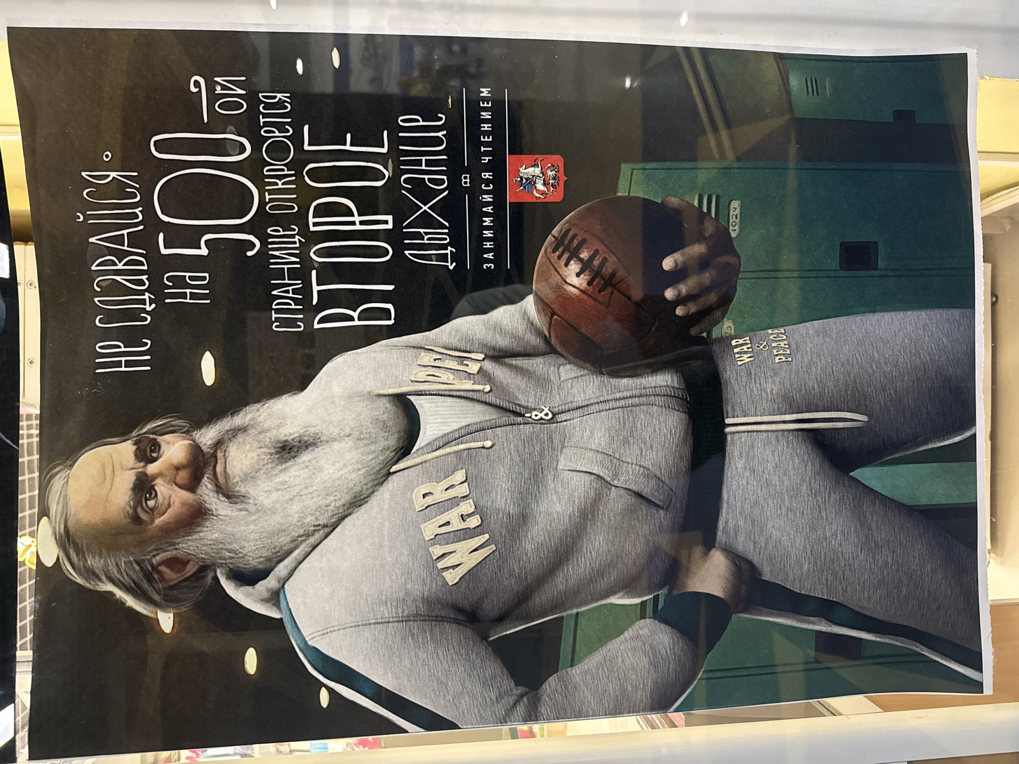 The image is a poster featuring a stylized illustration of an old man with a long white beard and hair, wearing a grey hooded sweatshirt with "WAR & PEACE" emblazoned on the chest and matching sweatpants. He is holding a vintage-style American football. The text on the poster reads in Russian: "Не сдавайся. На 500-ой странице откроется второе дыхание. Занимайся чтением." This translates to "Don't give up. On the 500th page, a second wind will open. Engage in reading." Below this text, there is a red emblem with a white horse and rider. The background is dark with some yellow circular lights. The poster appears to be displayed in a public space, possibly a library or an educational institution, given the theme of reading and the presence of lockers in the background. There is no indication of the specific location within the city of Votkinsk, Russia.