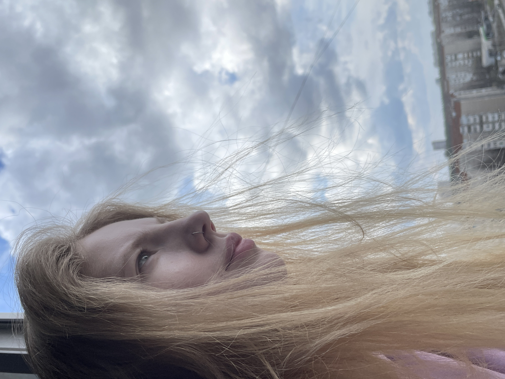 The image is a close-up, low-angle photograph of a young person's head and hair, captured with the camera rotated. The person, who has long blonde hair, is looking upwards towards the sky. Their face is visible from the nose up, revealing a septum nose ring. The voluminous blonde hair spreads across the bottom half and extends towards the right side of the frame, with individual strands distinctly visible against the background.
The background primarily features a bright, cloudy sky with patches of blue, indicating it is daytime. On the far right, a tall, multi-story building with numerous windows is partially visible against the sky. The building appears somewhat soft-focused, suggesting it might be viewed through a window or from a distance in an urban setting, possibly Izhevsk, Russia. The overall impression is one of quiet contemplation or observation of the outdoors. No specific activity beyond looking up is discernible, and no text is visible.