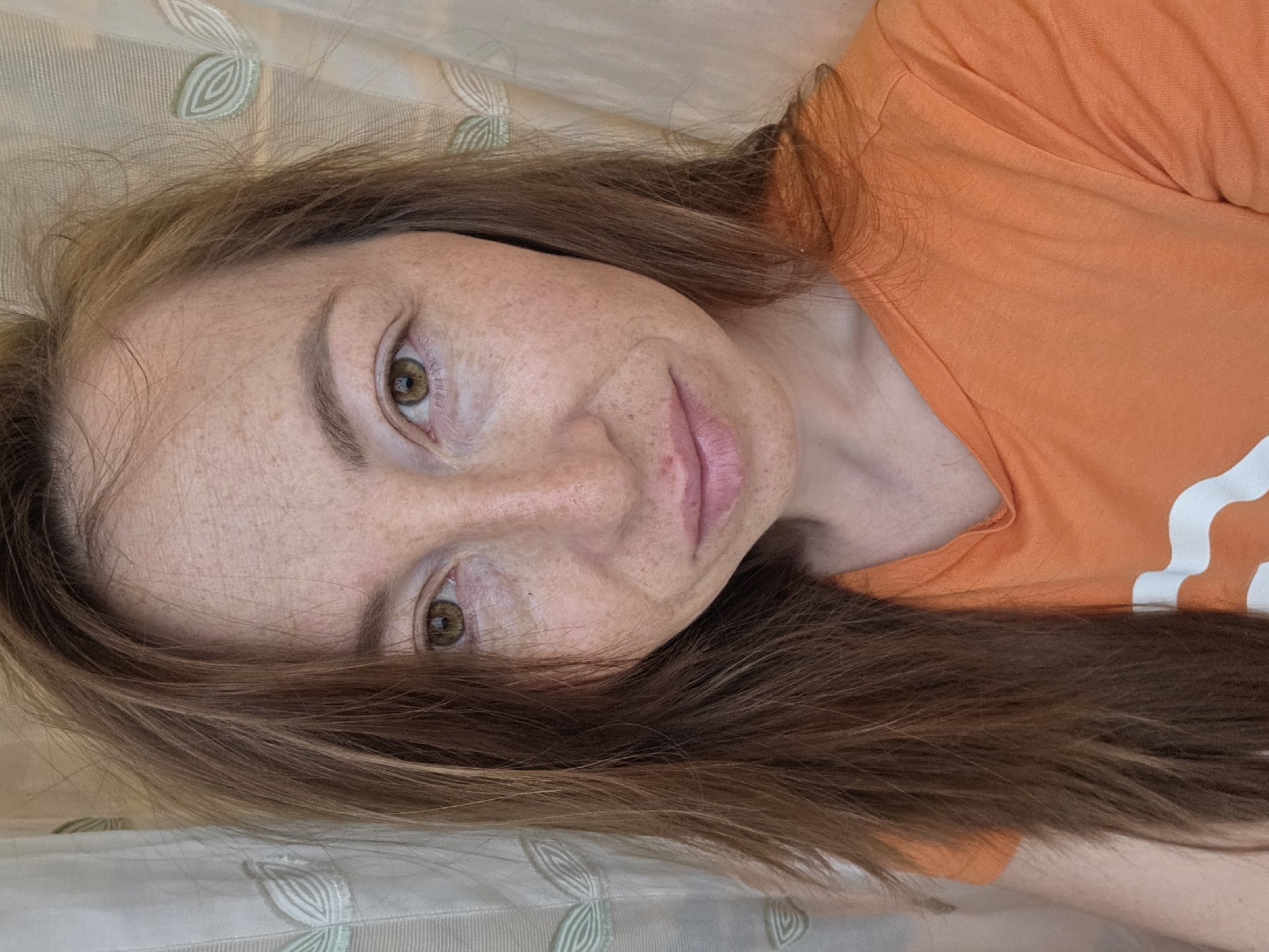 The image is a close-up, half-portrait shot of a person, likely female, lying down. The image is rotated 90 degrees counter-clockwise, with the person's head positioned towards the top left and their body extending to the bottom right.
**Subjects:** The central subject is a person with fair skin, notably covered in numerous freckles across their nose, cheeks, and forehead. They have long, light brown hair with subtle lighter highlights, which cascades around their face and rests on the bedding. Their eyes are a light brown or hazel color, looking directly towards the viewer. Their eyebrows are well-defined and light brown. Their lips are naturally pink and slightly parted.
**Activity or event taking place:** The person appears to be relaxing or lying down, possibly having taken a selfie.
**Notable details:** The person is wearing a bright orange t-shirt. A partial white graphic or text is visible on the right side of the shirt, appearing as a curved, thick white line. The background consists of light-colored bedding, possibly a pillow or sheet, featuring a subtle pattern of light green or beige, stylized leaf-like motifs. The lighting is soft and natural, suggesting an indoor setting during the daytime. The person's expression is neutral and calm.
**Visible text:** A partial white graphic on the orange t-shirt, resembling a curved white shape.