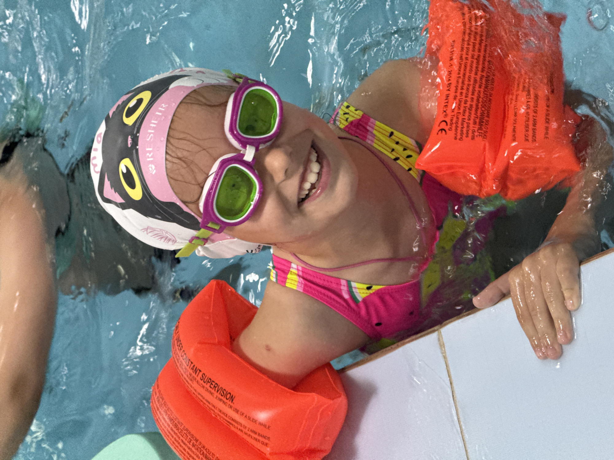 This is a close-up, overhead shot of a young girl in a swimming pool, wearing a pink and black cat-themed swim cap and purple goggles. She is smiling broadly, showing her teeth, and her head is tilted back, with her face close to the camera. She is wearing a brightly colored bikini with a yellow, pink, and black pattern. Orange arm floaties are on her arms, and one is also around her neck. The water is blue and appears somewhat murky, with ripples on the surface. The edge of the pool, made of white tiles, is visible in the foreground and right side of the image. Text on the arm floaties is partially visible and appears to be safety instructions in multiple languages, including "UNDER CONSTANT SUPERVISION" and "CE". The setting is clearly a swimming pool, likely an indoor one given the lighting. The girl's expression conveys happiness and enjoyment. The image captures a moment of fun and play in the water.