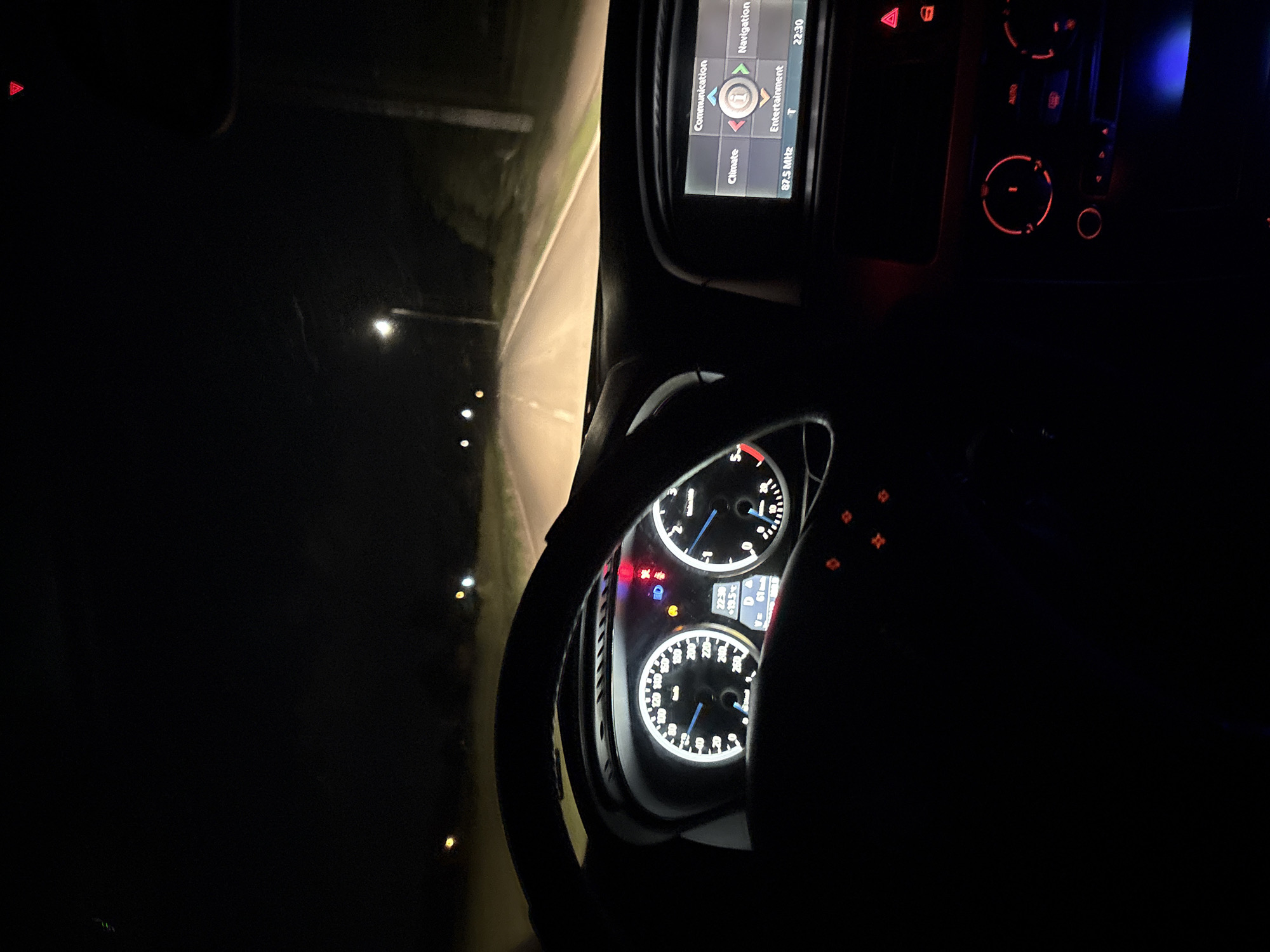 The image depicts a view from inside a car driving at night. The dashboard and instrument panel are visible, showing the car's speed and other information. The radio is tuned to 87.5 MHz and the car's GPS is active. Outside, the road is illuminated by streetlights.