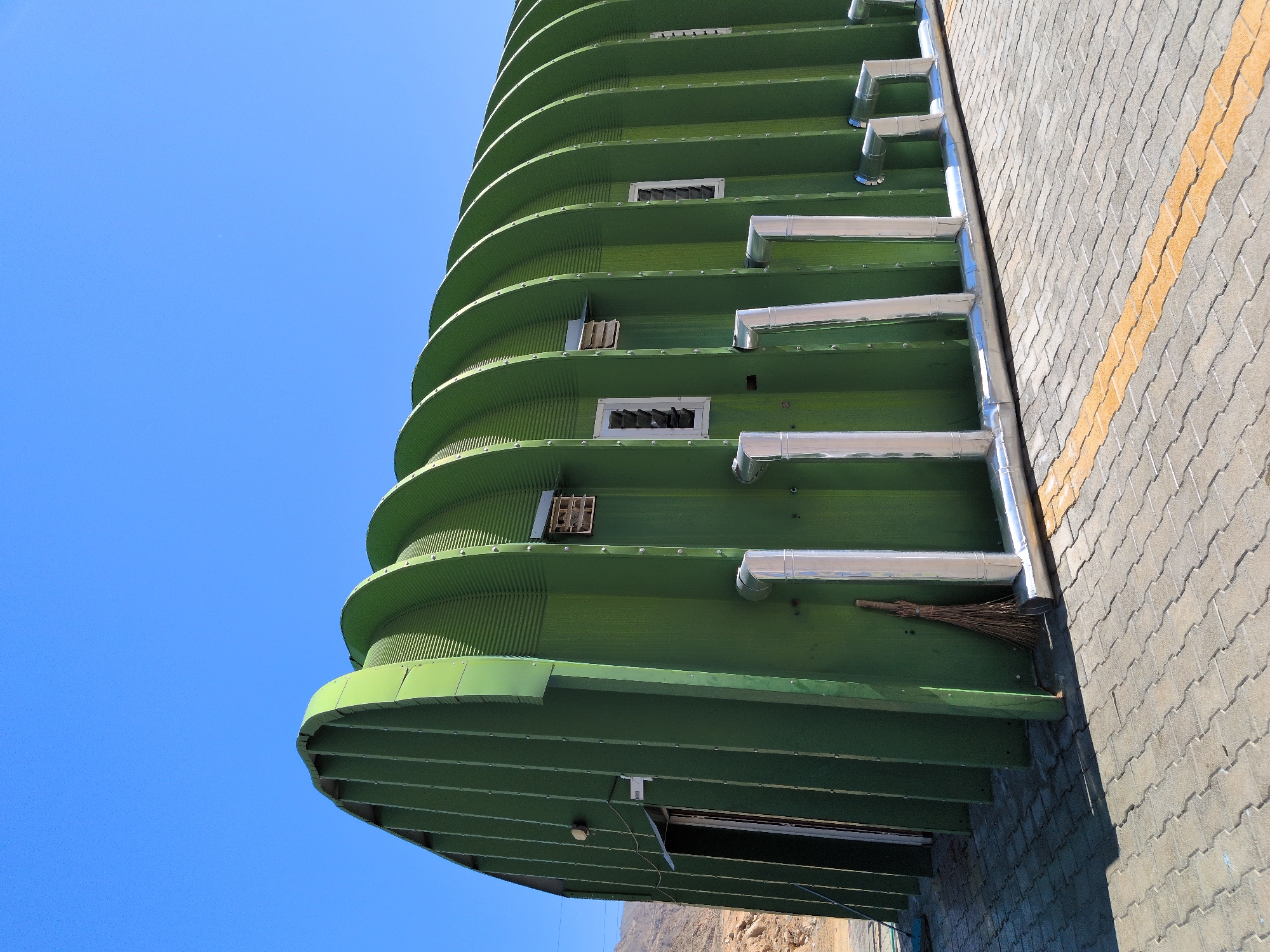 The image shows a long, green, Quonset hut-style building under a clear blue sky. The building is constructed from corrugated metal panels and has a series of arched ribs along its length. There are several windows with louvers and what appear to be air vents on the side of the building. Silver-colored pipes run along the base of the structure. A broom leans against the building near the front entrance, which is slightly ajar. The ground in front of the building is paved with interlocking concrete pavers, with a yellow stripe marking a boundary. The background shows a rocky, arid landscape with hills in the distance. The scene is illuminated by bright sunlight, casting shadows on the ground. The location is identified as Chumkar, India. There are no people visible in the image.