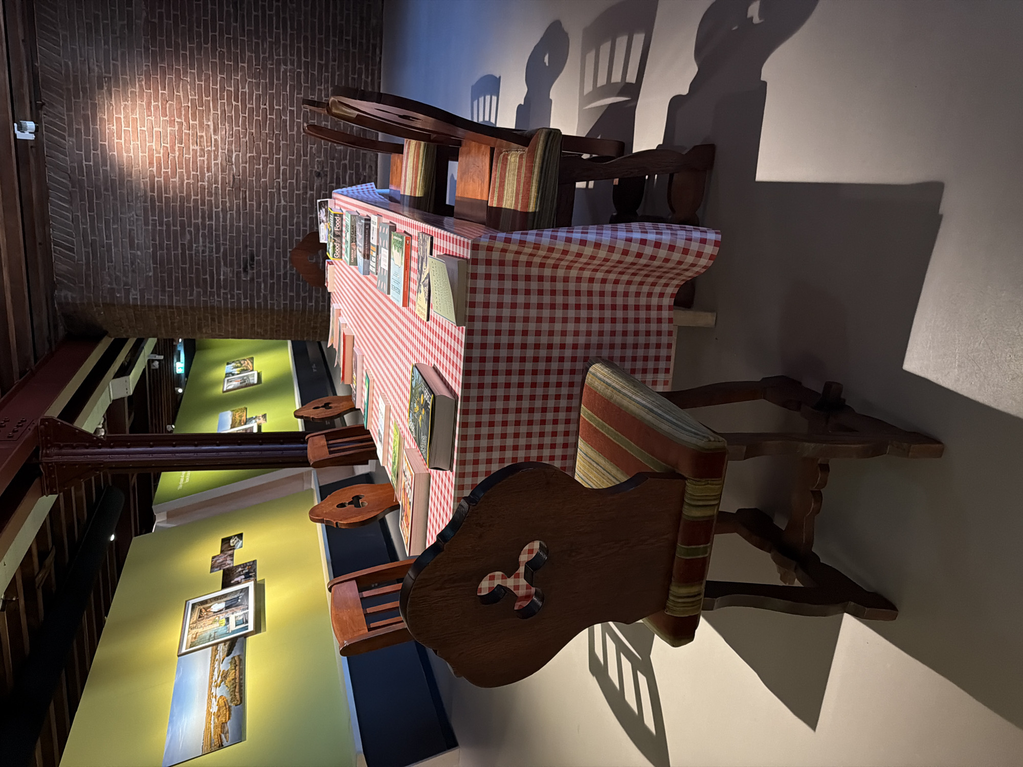 The image shows a long table covered with a red and white checkered tablecloth, set in a room with exposed brick walls and wooden beams. The table is laden with books, some stacked and others open. Around the table are several wooden chairs with upholstered seats. The room appears to be an exhibition space, with framed photographs displayed on a lime green wall in the background. One photograph depicts a scenic landscape with water and reeds, and another shows a person standing in an outdoor setting. The lighting in the room is dramatic, casting long shadows from the chairs onto the floor. This scene is located within the Maritime Museum in Rotterdam, Netherlands, which houses a diverse collection of artifacts and exhibits related to seafaring history and maritime culture.