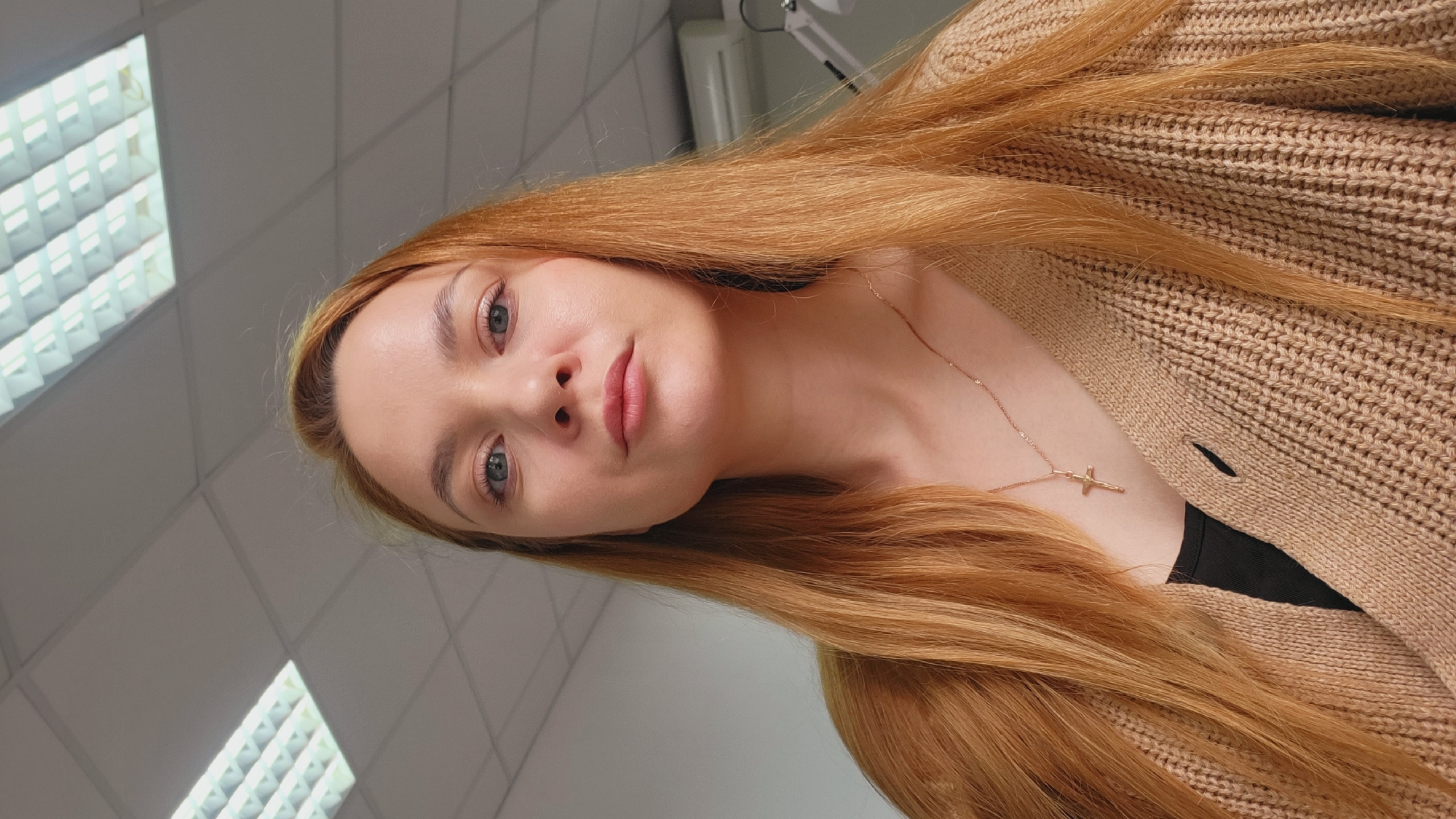 This close-up portrait features a young woman with long, straight, reddish-blonde hair, which flows over her shoulders and chest. She gazes directly at the camera with light-colored eyes and a neutral, calm expression, her lips slightly parted. She is wearing a light brown, chunky knit cardigan over a black top. A delicate gold-colored chain necklace with a small cross pendant rests on her décolletage.
The setting is indoors, with a light-colored ceiling composed of square tiles visible in the background. Several bright, recessed fluorescent light fixtures are illuminated, suggesting an office, commercial building, or institutional environment. A piece of white equipment is partially visible in the upper right of the frame. The artificial lighting makes the time of day indiscernible. The woman appears to be posing for a photograph. No text is visible in the image. The original image file is oriented 90 degrees clockwise.