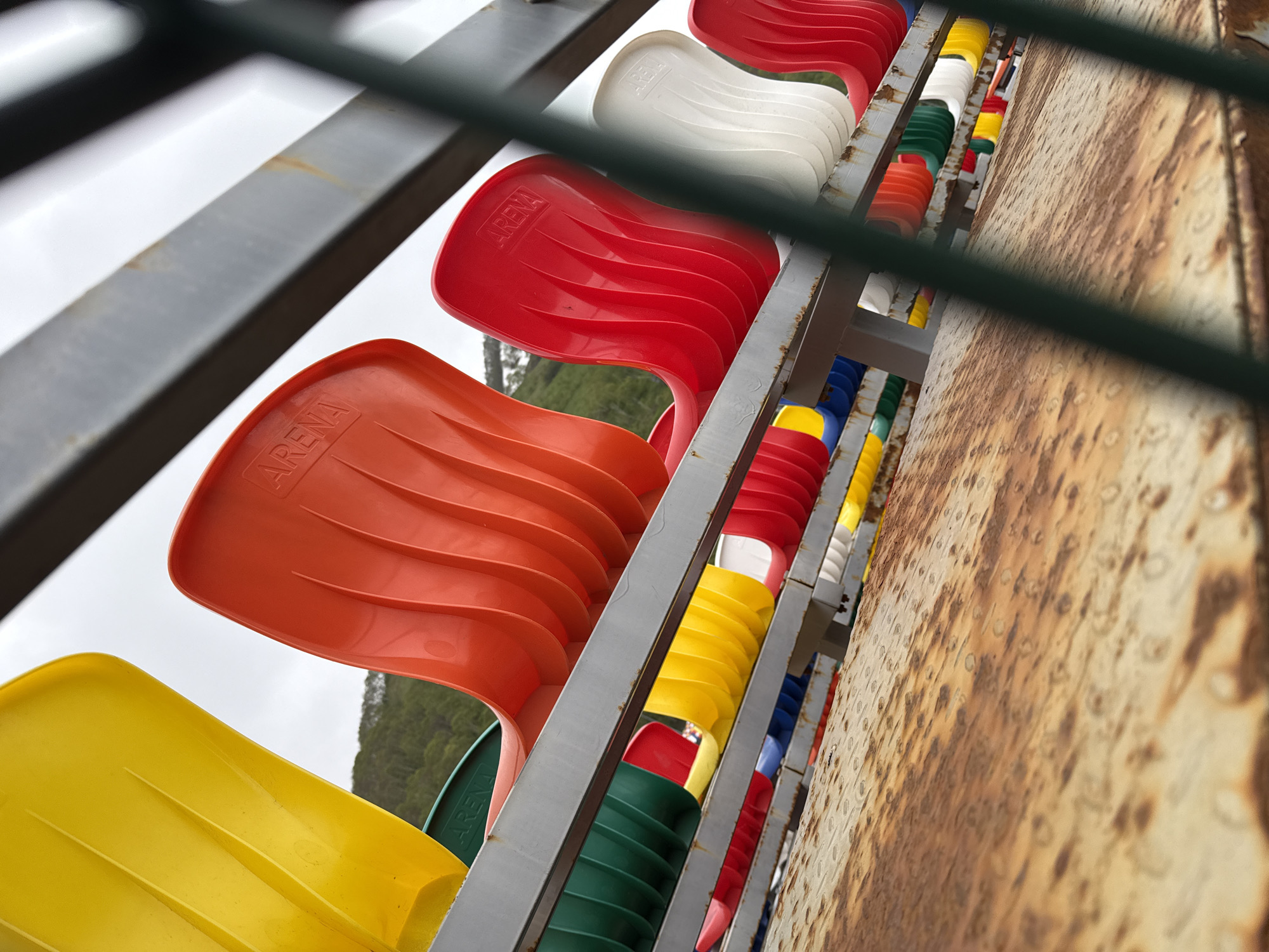 A low-angle shot of colorful stadium seats in Petropavlovsk-Kamchatsky, Russia. The seats are arranged in rows on metal bleachers, with some of the seats being red, yellow, green, and white. The metal structure supporting the seats is rusted. In the background, a green, forested hillside is visible under a cloudy sky. A dark green fence or railing is in the foreground, partially obscuring the view. The word "ARENA" is embossed on some of the red seats. The overall scene suggests an outdoor sports venue or amphitheater.