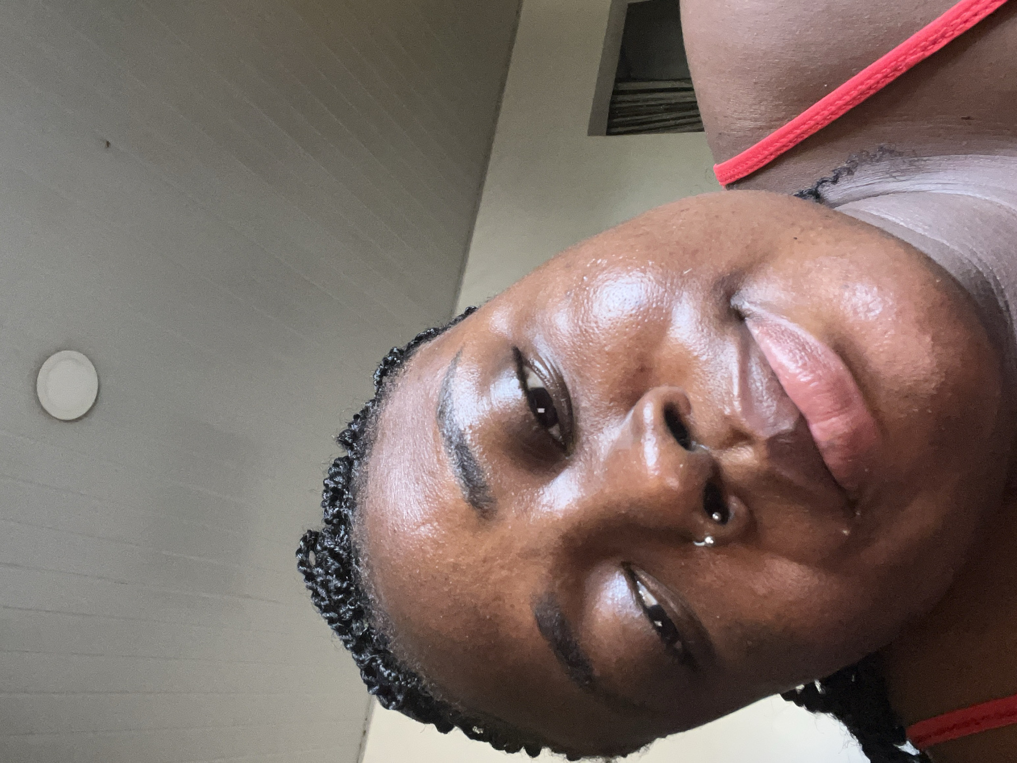 The media file is a low-angle, close-up, tilted selfie of a young Black woman. She is wearing a coral-colored bikini top and appears to be outdoors, as indicated by the natural light and the presence of building exterior details. The background suggests an urban or suburban setting, possibly a patio or balcony, with a neutral-colored wall and a glimpse of what might be a window frame.
The woman's face is the central focus, with her skin appearing dewy and possibly sweaty, suggesting warm weather. She has braided hair, with some of the braids framing her face. She is wearing a silver nose ring. Her expression is somewhat subdued, with her eyes looking slightly away from the camera and her mouth partially open. The lighting suggests it is daytime. The overall impression is a candid moment captured in a relaxed, possibly humid, environment in Abuja, Nigeria. There is no visible text in the image.