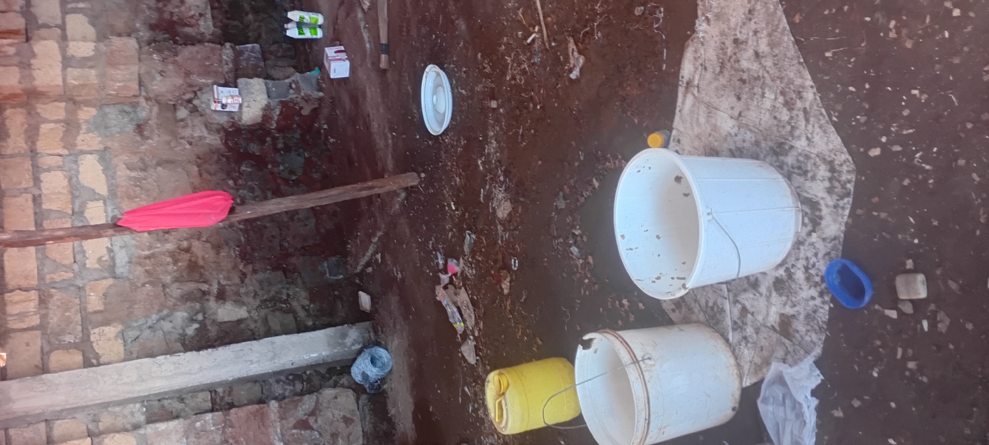 The image depicts an interior scene within a structure characterized by rough, unfinished brick walls. A wooden post stands vertically in the center, adorned with a folded bright pink fabric. Various objects are scattered on the dirt floor, including two white buckets, one yellow plastic jerrycan, and a white plate-like object. There are also some debris, including plastic packaging, bottles, and what appears to be a roll of wire fencing in the background on the left. In the upper right corner, on a raised brick surface, are two bottles of cleaning liquid and a small white box. The overall environment appears to be a storage or work area, possibly in a semi-rural or undeveloped setting. The location is identified as Kijabe Ward, Kenya.