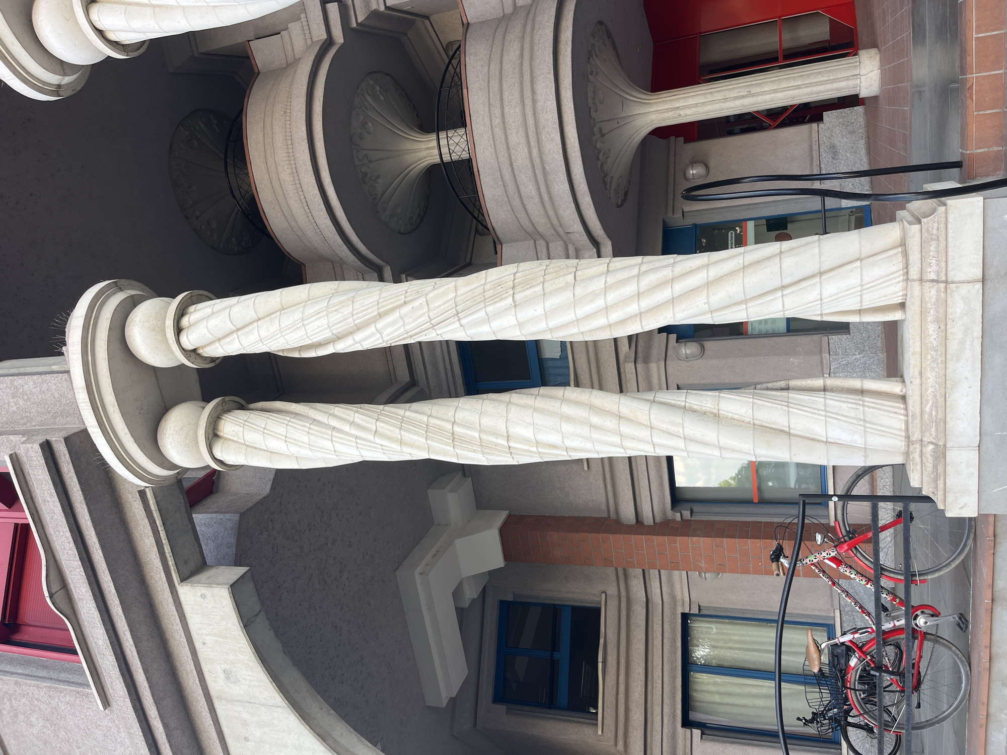 The image shows the exterior of a building with an unusual architectural design. Two large, white, twisted columns dominate the foreground. Behind them, the building features multiple levels with rounded balconies, red window frames, and decorative circular elements under the balconies. There are also windows with blue frames and some exposed red brickwork. In front of the building, two bicycles are parked. The overall impression is one of modern, somewhat eccentric architecture. The location is confirmed to be Geneva, Switzerland.