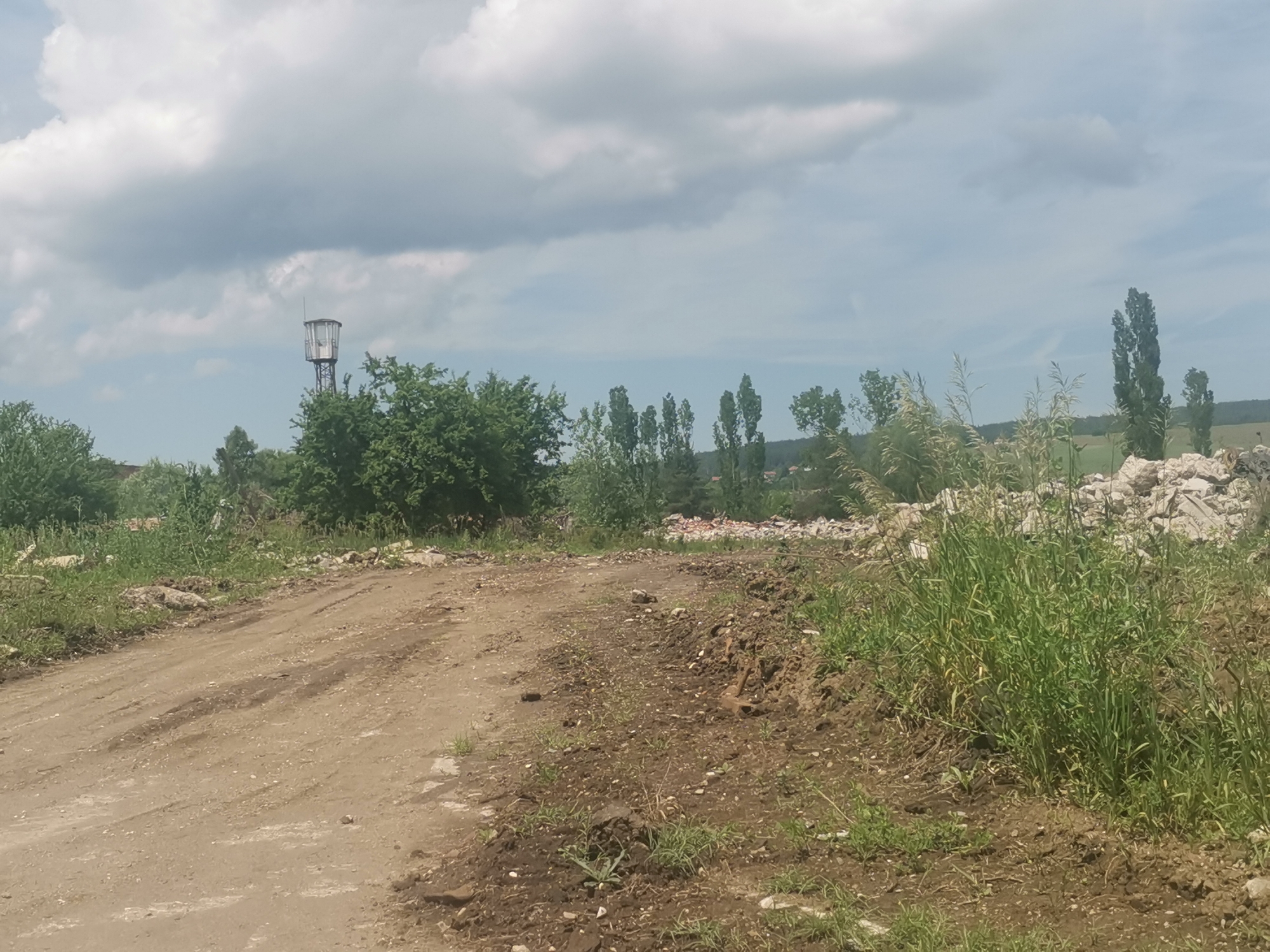 This image depicts a rural or semi-rural landscape in Zhivkovo, Bulgaria, under an overcast sky. A wide, light-brown dirt road curves through the foreground and mid-ground, showing tire tracks and small stones. On either side of the road, the ground is a mix of exposed dark soil and vibrant green grass and weeds.
In the mid-ground, to the right, large piles of light-colored rubble or construction debris are visible, partially covered by tall, wild grass. A line of slender, tall trees, possibly poplars, stretches across the mid-ground and into the background on the right. On the left side of the frame, a dense mass of green deciduous trees and bushes forms a buffer.
A notable object is a tall, metal water tower with a distinct, enclosed cabin-like structure at its apex, positioned on the left, rising above the tree line. In the far distance, hints of a landscape with trees and potentially distant buildings with reddish roofs can be seen on the horizon.
The sky is largely covered by large, fluffy grey and white clouds, indicating an overcast but bright daytime. There are no people visible in the scene, and no discernible text. The overall impression is one of a quiet, somewhat wild or undeveloped area, with elements of utility (water tower) and potential past or ongoing activity (rubble, dirt road).