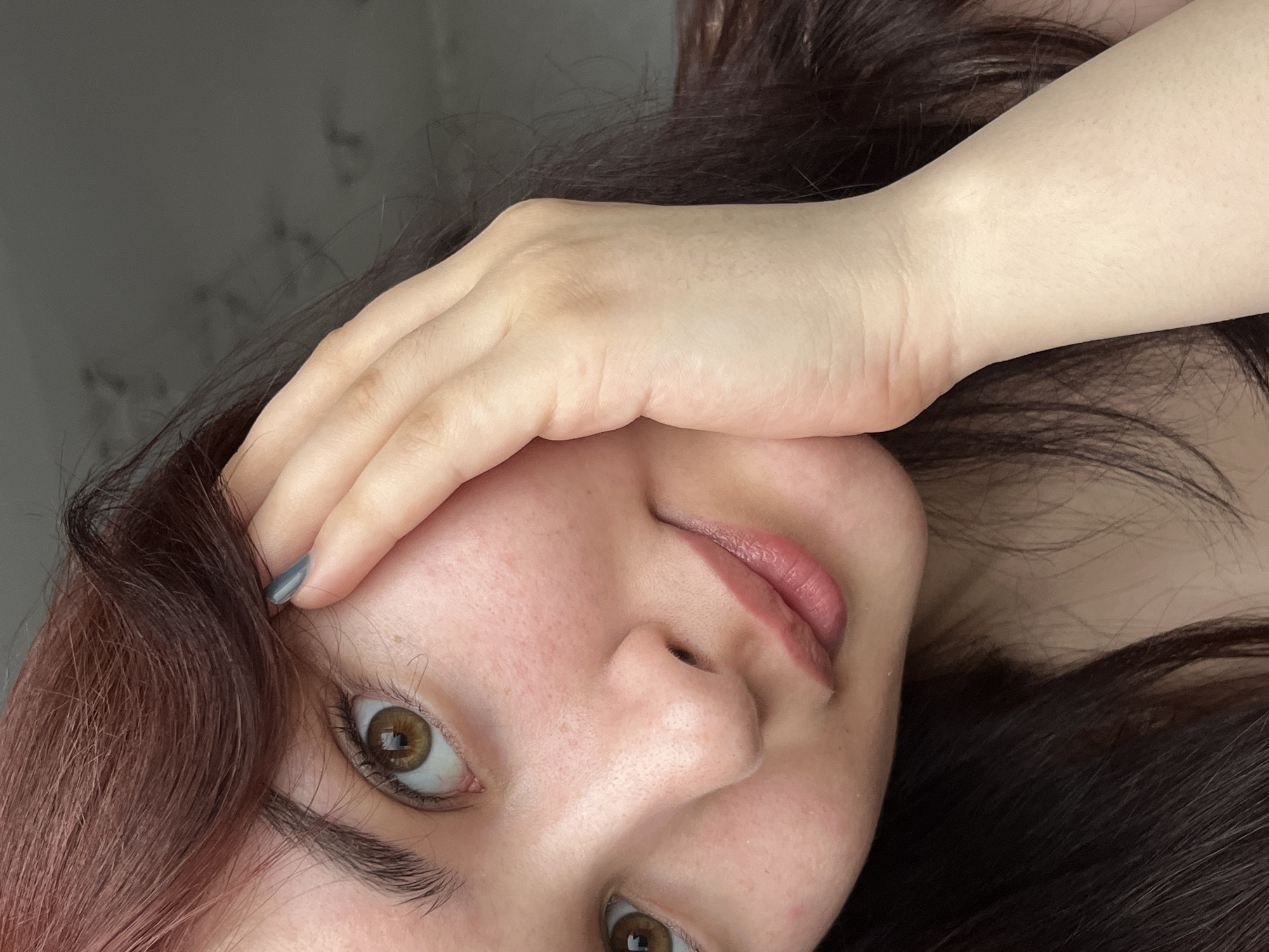 A close-up, slightly rotated image focuses on a woman's face and upper hand. Her face is characterized by fair skin, brown eyes, and pink lips. She has brown hair, with hints of red or auburn tones. Her hand is positioned near her forehead, partially obscuring her face. Her fingernails are painted a grey color. The background appears to be a plain, light-colored wall or surface. There is no indication of Neftekamsk, Russia or any other city location.