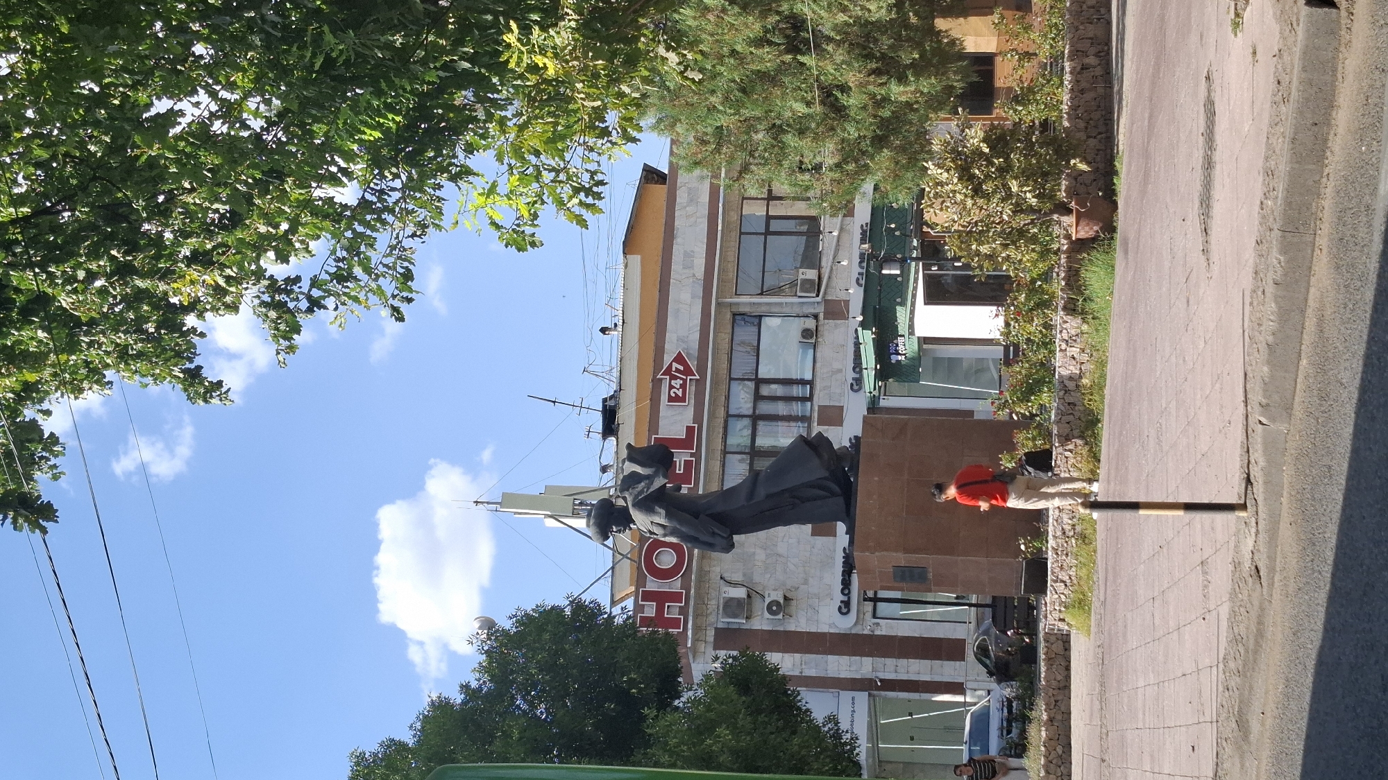 The image captures a sunlit urban scene, likely in Shymkent, Kazakhstan, featuring a prominent statue in the foreground. The statue, a dark, monolithic figure with flowing garments and a head covering, stands on a reddish-brown stone pedestal. Behind the statue is a building with a large "HOTEL" sign in red lettering. The hotel appears to be a multi-story structure with a facade of marble-like panels and a darker band near the roof. A "24/7" sign with an arrow is visible to the right of the "HOTEL" sign. Several air conditioning units are mounted on the building.
In front of the hotel, a cafe or shop with a green awning is visible, displaying "PRO COFFEE" signage. The area around the statue and building is paved with light-colored square tiles, interspersed with grassy areas and low stone walls. A person wearing a red shirt and beige pants is standing on the tiled area, facing away from the camera, holding a mobile phone. To the left, two cars are parked on the street, and a woman in a striped shirt and dark pants is standing near them. Lush green trees, some with broad leaves and others with needle-like foliage, frame the scene, with power lines visible against the bright blue sky dotted with white clouds. The overall impression is of a public space with a blend of architectural elements and natural greenery.