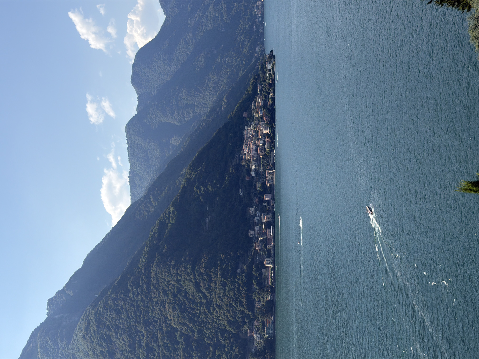 The image depicts a wide view of Lake Como, with the town of Pognana Lario situated along its shoreline, nestled at the base of steep, tree-covered mountains. The water is a deep blue, with a few small boats creating white wakes. The town itself is a cluster of buildings with red-tiled roofs, seemingly clinging to the mountainside. The sky is a bright blue, with a few scattered white clouds. The overall impression is one of a serene, picturesque landscape.