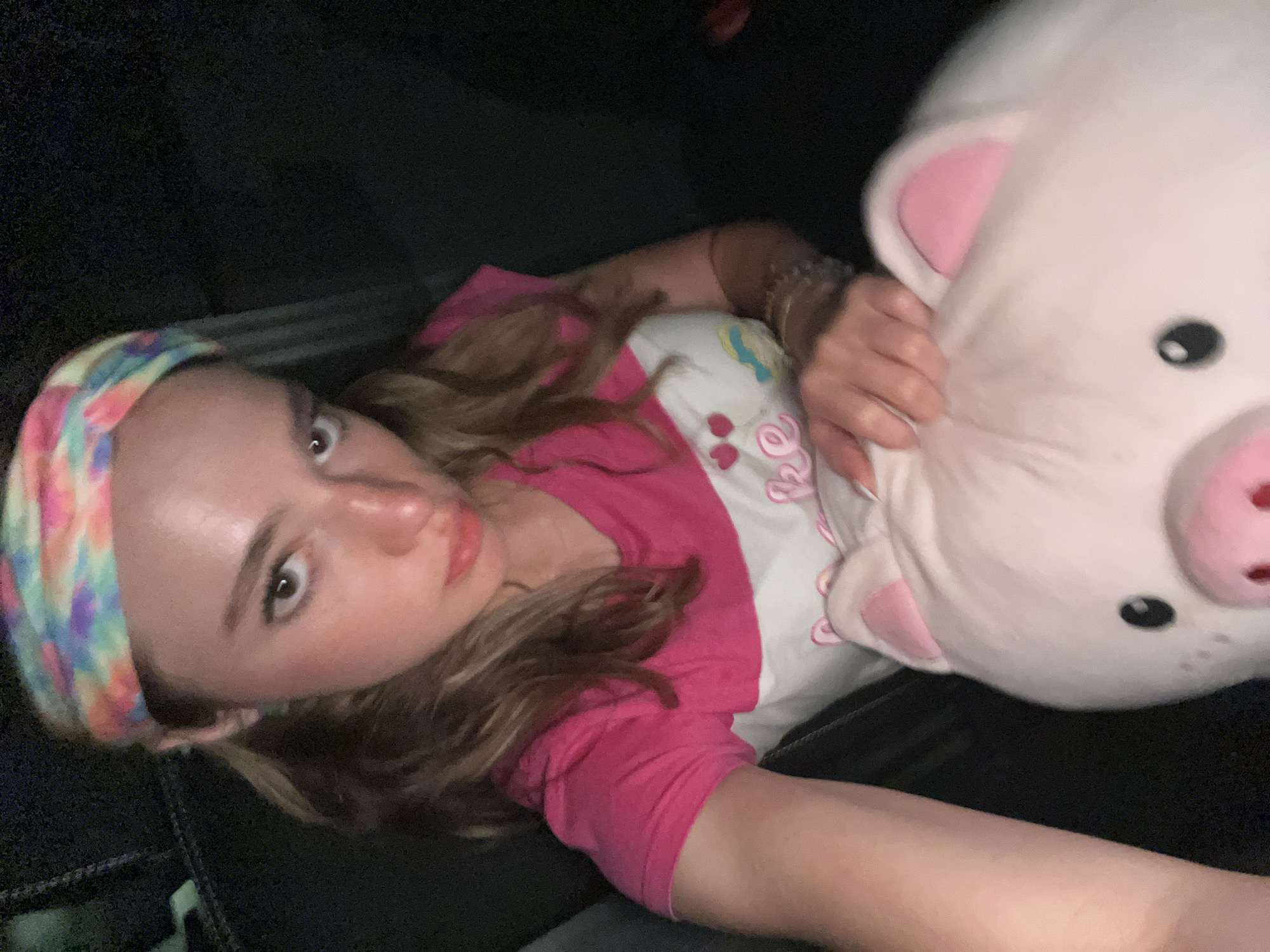 A young woman lies on a dark, textured surface, possibly fabric, while holding a large, plush pig toy. She wears a pink t-shirt over a white shirt with small cartoon graphics, and a colorful, patterned headband. She is looking up and towards the camera. The setting is dark. There is no location information.