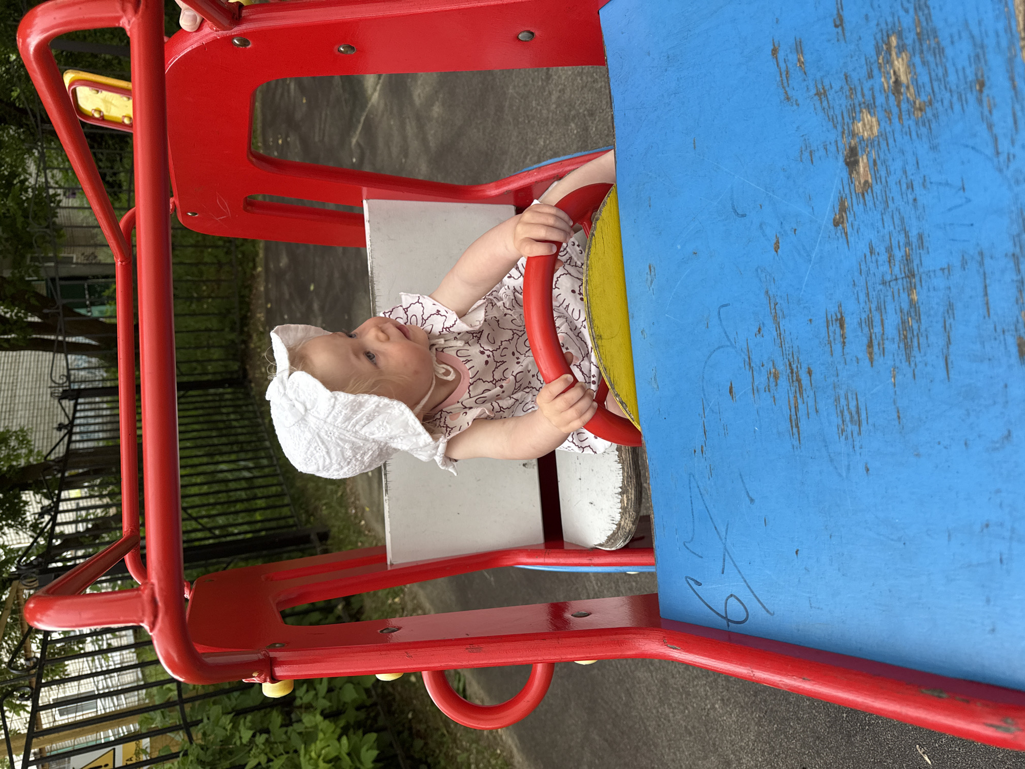 A young Caucasian child wearing a white sun hat and a pink dress with a white bear pattern sits in the driver's seat of a bright red and blue play vehicle, likely a toy truck or car. The child is looking to the right of the frame, with their hands on a red steering wheel. The play structure appears to be on a paved surface, possibly a playground or park, with a black metal fence and greenery visible in the background. There are faint scratches and markings on the blue part of the play vehicle. The image was taken outdoors on a cloudy day. The location is identified as Vladimir, Russia.
