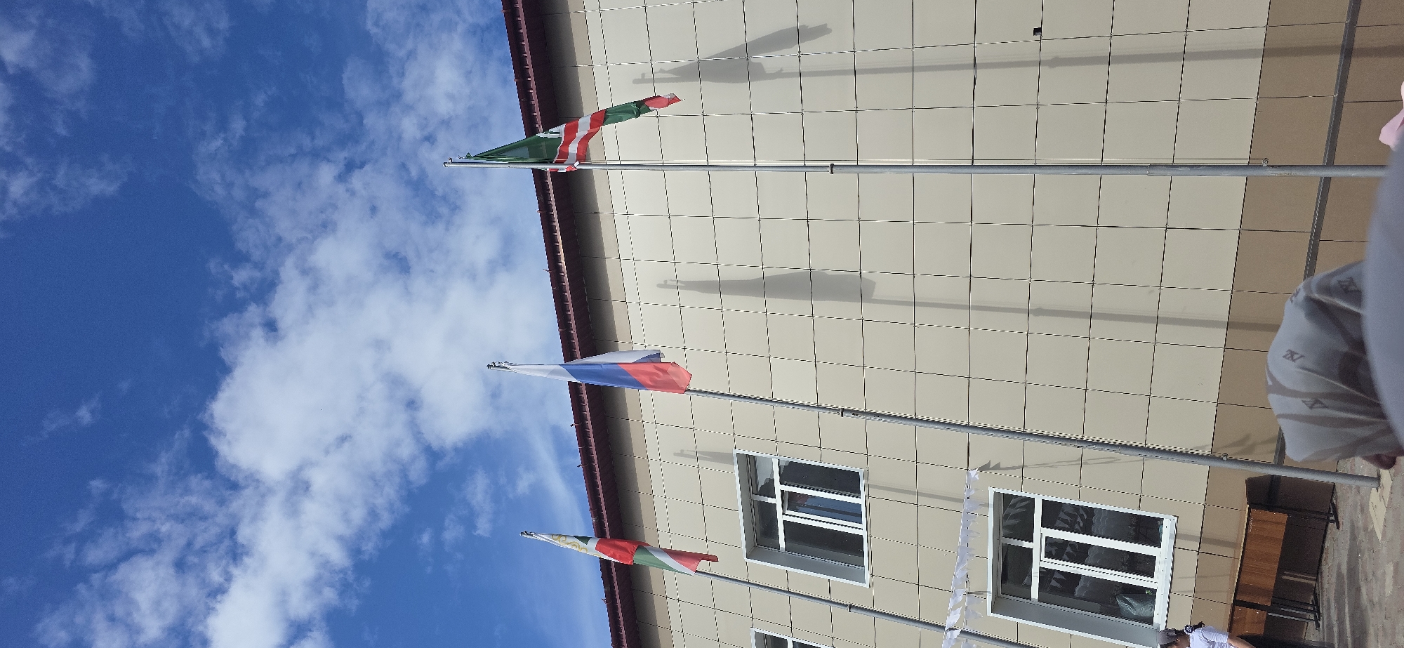 Here is a forensic description of the image:
The image captures a low-angle view of a building facade under a partly cloudy blue sky. Three flags are prominently displayed on poles attached to the building. The top flag features a green and red striped design. The middle flag displays the colors white, blue, and red, arranged horizontally. The bottom flag mirrors the design of the top flag. The building has a light-colored exterior with a grid-like pattern and several windows, all appearing to be closed. The building exterior also reflects the shadows of the flag poles. The camera angle creates a perspective where the building seems to tower over the viewer. A person's side is partially visible in the frame with their face obscured. The photograph seems to have been taken outdoors during daylight hours, based on the presence of natural light and the appearance of the sky. There is little evidence to identify a specific location.