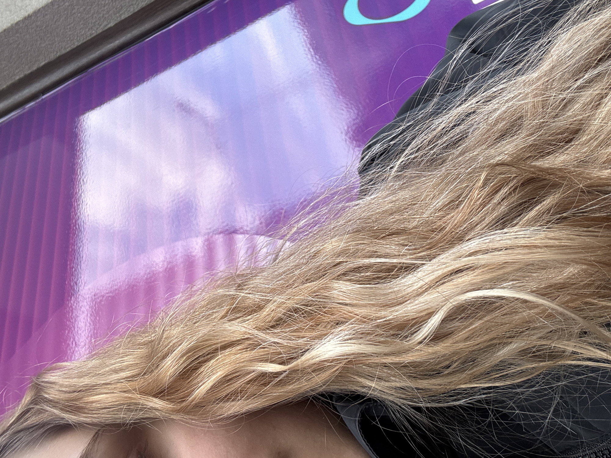 The image presents a close-up, abstract view dominated by a person's blonde hair against a reflective, purple background. The upper two-thirds of the frame are filled with a glossy, vibrant purple surface, which features subtle vertical striped patterns. This surface highly reflects the ambient light, showing what appears to be clouds or the sky in soft, diffused white and light blue tones. In the top right corner of this purple surface, a segment of a light blue, rounded graphic or letter is partially visible.
In the lower portion of the image, a significant volume of wavy, golden-blonde hair extends across the frame. The hair appears natural and slightly dishevelled, with individual strands catching the light, highlighting variations in tone. Some darker hair roots are discernible beneath the lighter blonde. Below and to the right of the main mass of hair, a section of dark, possibly black, fabric is visible, likely part of clothing or an accessory, offering a stark contrast to the bright hair. A very small portion of the person's face, including the forehead, the bridge of the nose, and the area around an eye, is faintly visible along the bottom edge, partially obscured by the hair.
The scene takes place outdoors during the daytime, as indicated by the clear reflections on the glossy purple surface. No specific activity is evident beyond the composition of the close-up shot, possibly taken from a slightly elevated perspective or a selfie. There is no decipherable text in the image. The location context for this media is Montana, Bulgaria.