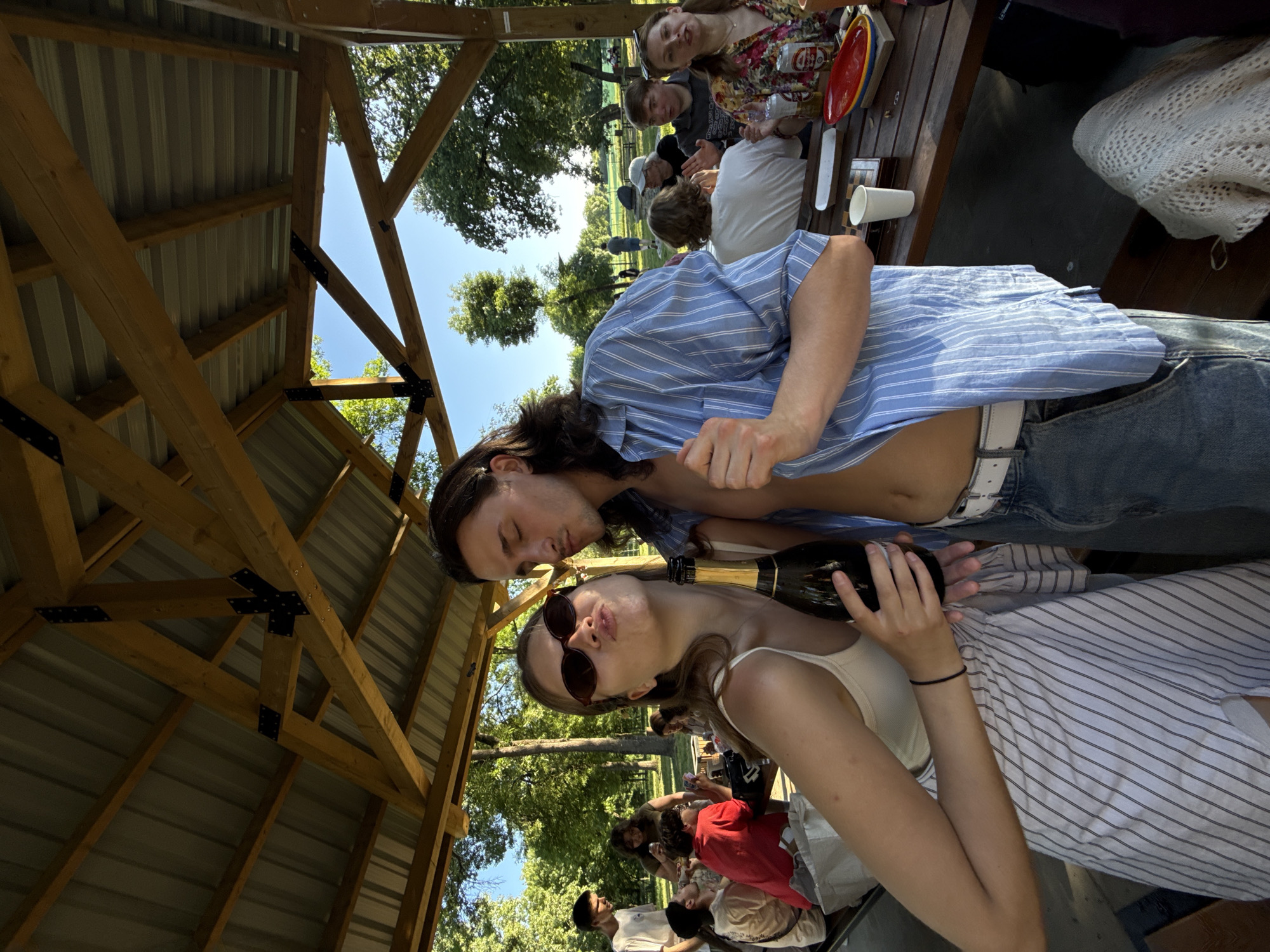 The scene depicts a group gathered outdoors, likely at a park or recreational area, under a wooden-roofed structure, possibly a gazebo or picnic shelter. A woman with sunglasses holds a bottle, seemingly a champagne or sparkling wine bottle, while a man stands closely behind her. Other individuals are visible in the background, seated around a table with various items, including plates and cups, suggesting a casual social gathering or picnic. The trees in the background indicate the presence of nature. However, the image doesn't provide enough detail to definitively confirm the location as Belgrade, Serbia, or to determine the specific identities or relationships of those present.