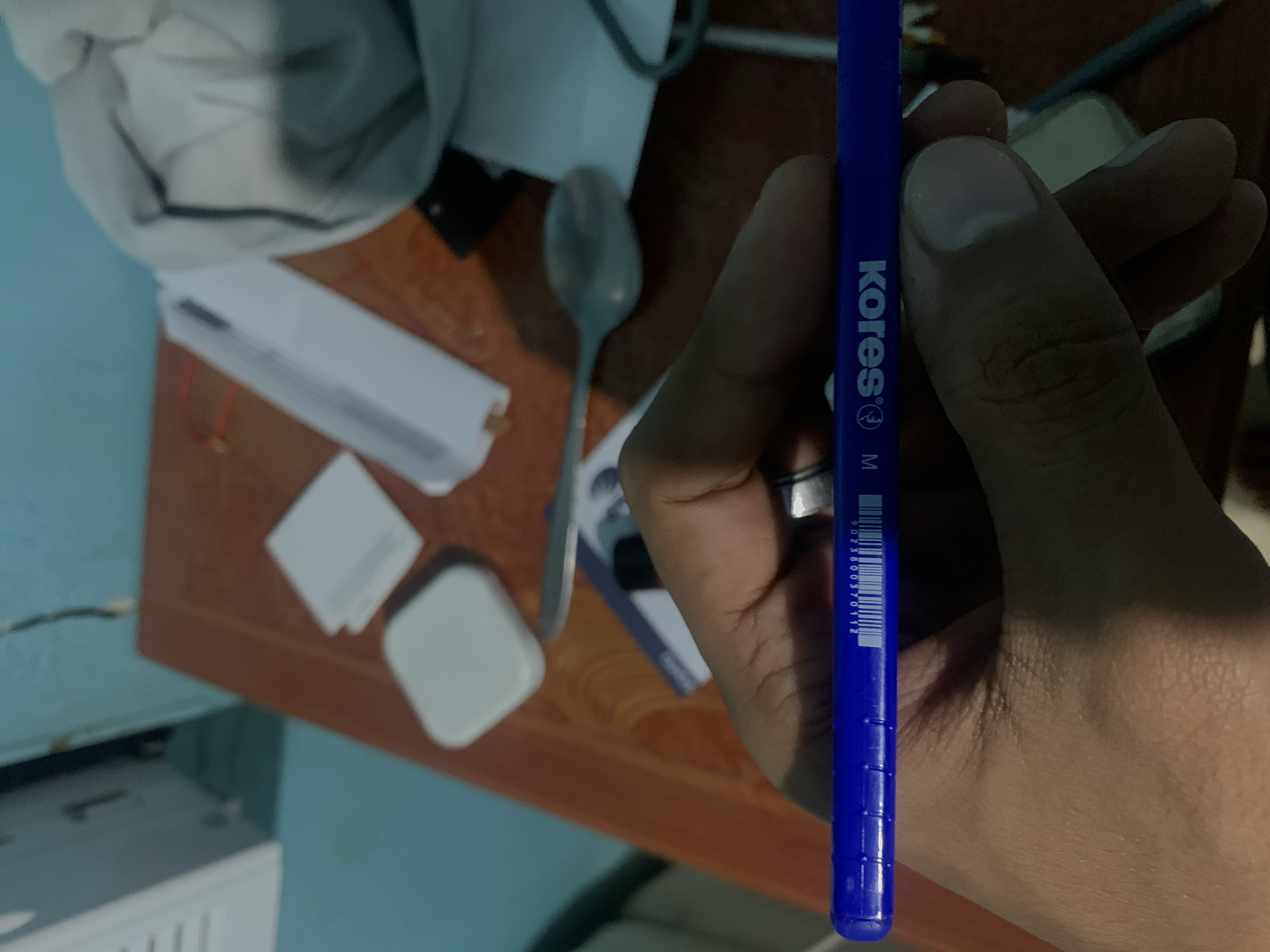 The image captures an indoor scene in Santa Ana, Venezuela, focusing on a person's hand holding a blue pen. The hand, wearing a ring on one finger, grips the pen vertically in the right side of the frame. The pen features the brand name "Kores® M" printed in white, along with a barcode displaying the numbers "9023800370112".
The background reveals a wooden table surface where several items are scattered. These include a silver spoon, a white rectangular case or container, a small white square object, and another white object with rounded corners. In the upper left, soft white and light blue fabric, possibly clothing, is visible. The far left background shows a light blue wall, while a partially visible white electronic device (like a printer or speaker) can be seen on the bottom left. The lighting is consistent with indoor ambient light. The activity depicted is simply the act of holding the pen, potentially in preparation for writing or as an object of focus. No specific emotions are discernible.
