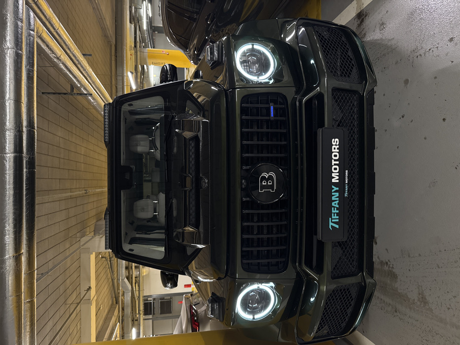 A dark-colored Brabus G-Wagen, a high-performance modified Mercedes-Benz G-Class, is prominently featured in this image, likely situated in an indoor parking garage or industrial-style showroom. The vehicle is captured from a front-three-quarter perspective, with its driver's side door open, offering a glimpse of its light-colored, possibly white or cream, interior seats.
The front of the SUV displays a bold "B" logo, indicative of Brabus, centered on its distinct matte black grille. The round headlights are illuminated with bright LED rings, and the vehicle features an aggressive, muscular front bumper design with large air intakes. A black license plate holder is attached to the front grille, clearly displaying the text "TIFFANY MOTORS" in a light blue-green color, with the same text repeated in a smaller font below it.
The background reveals architectural elements typical of an indoor facility. Beige brick walls are visible, with insulated silver pipes running horizontally along them. Concrete surfaces also make up parts of the walls and floor. Overhead, some yellow structural elements or large pipes can be seen. The artificial lighting suggests an enclosed space, and no people are visible in the frame. The scene is located in Warsaw, Poland, showcasing this luxury vehicle in a functional or display setting.