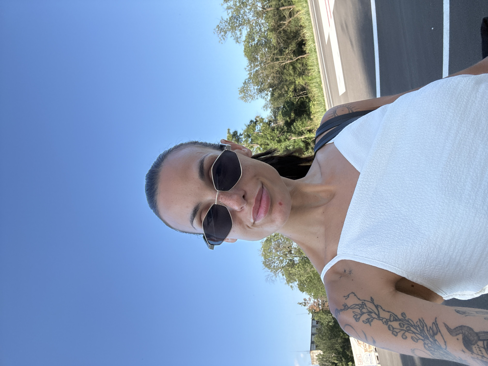 A woman is taking a selfie outdoors. She is wearing sunglasses and a white top. She has tattoos on her left arm. The background shows a clear blue sky, trees, and a road. The location is a street in Pleven, Bulgaria.