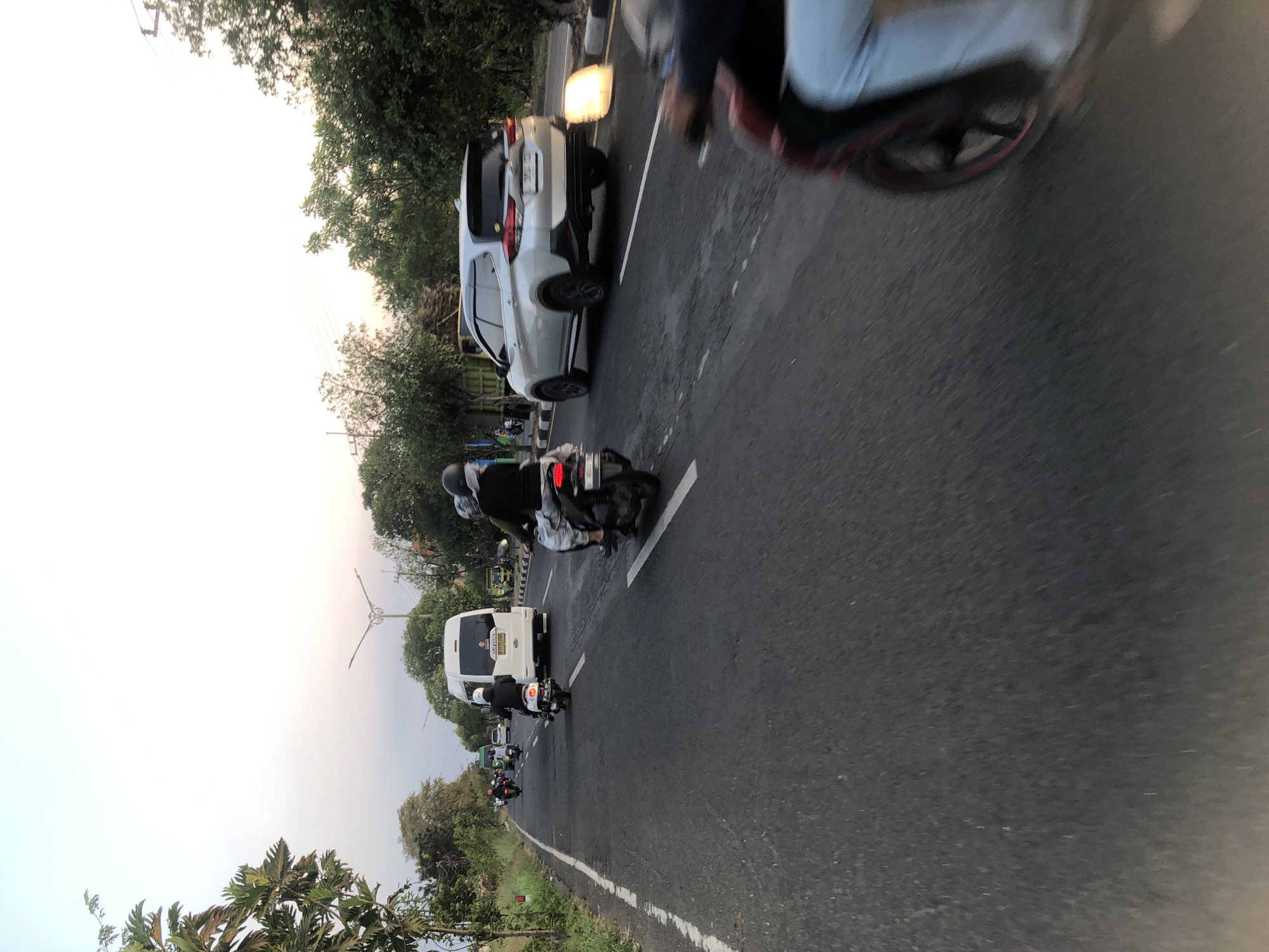 The image captures a street scene in Siring, Indonesia, during what appears to be late afternoon or early evening. The perspective is from a moving vehicle, indicated by the motion blur in the foreground and the road ahead.
The road is paved and has lane markings. On the right side of the road, a silver SUV is driving, and a bright yellow light source is visible near its rear bumper, possibly a street light or a hazard light. Beyond the SUV, there are trees and foliage lining the road.
Several motorcycles are also present on the road, with riders. One motorcycle in the foreground is heavily blurred, showing a person on it. Another motorcycle further ahead has a rider wearing a dark shirt and a white helmet. A white van is visible in the middle distance, driving in the same direction. In the background, more motorcycles and a green truck can be seen, along with more trees and buildings. The sky is a light, hazy blue, suggesting either a cloudy day or the sun setting. The overall impression is a typical road in a populated area of Indonesia, with active traffic.