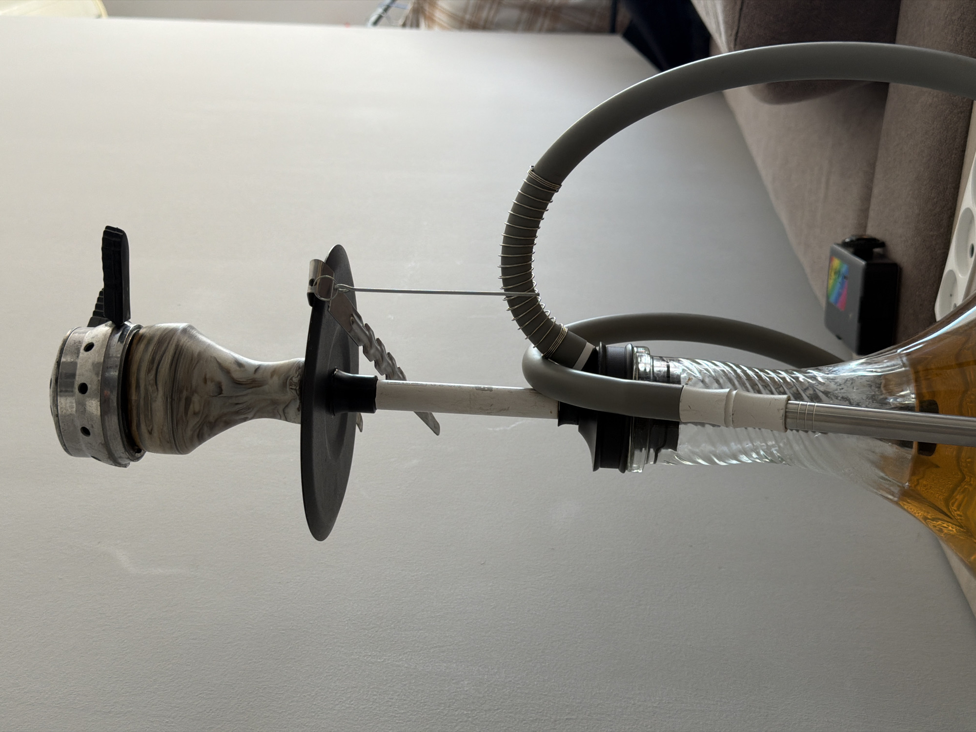 This is a close-up, medium shot of a hookah set up on a surface that appears to be a table or shelf, with a beige couch in the background. The hookah is made of metal and glass, with a patterned ceramic or resin bowl at the top. A grey hose extends from the side of the hookah. The liquid in the glass base is a golden-yellow color. A small black electronic device with a colorful, abstract pattern is visible on the couch. The wall behind the hookah is plain and textured. The overall lighting suggests an indoor environment, possibly a living room. There are no people or animals visible in the image.