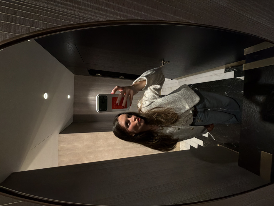 A woman stands in front of an oval mirror, capturing her reflection with her phone. She wears a cream-colored jacket over a frilly white shirt and jeans. The background shows a corridor with alternating black and white marble flooring, wood paneling, and recessed lighting. The top of the frame reflects the ceiling with what seems to be dark brown wood panels.