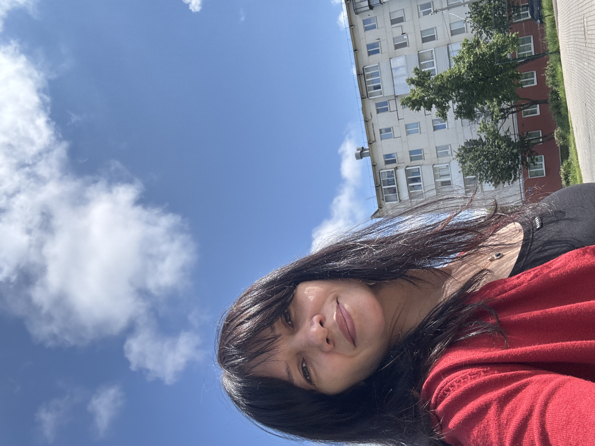 A woman with long, dark hair, wearing a red long-sleeved top over a black garment, takes a selfie outdoors under a bright blue sky. She has a subtle, pleasant smile on her face, with her dark hair framing her features. The background features a clear blue sky dotted with scattered white cumulus clouds, occupying the upper left portion of the image. On the right, a multi-story residential building with light-colored walls, numerous windows, and balconies is visible, partly obscured by green trees. A paved path or sidewalk runs along the far right edge of the frame, adjacent to the trees and building. The scene is brightly lit, indicating a sunny daytime. Faint white text, possibly "LIFTS," is visible on the black strap of her top. The location is Chaikovsky, Russia.