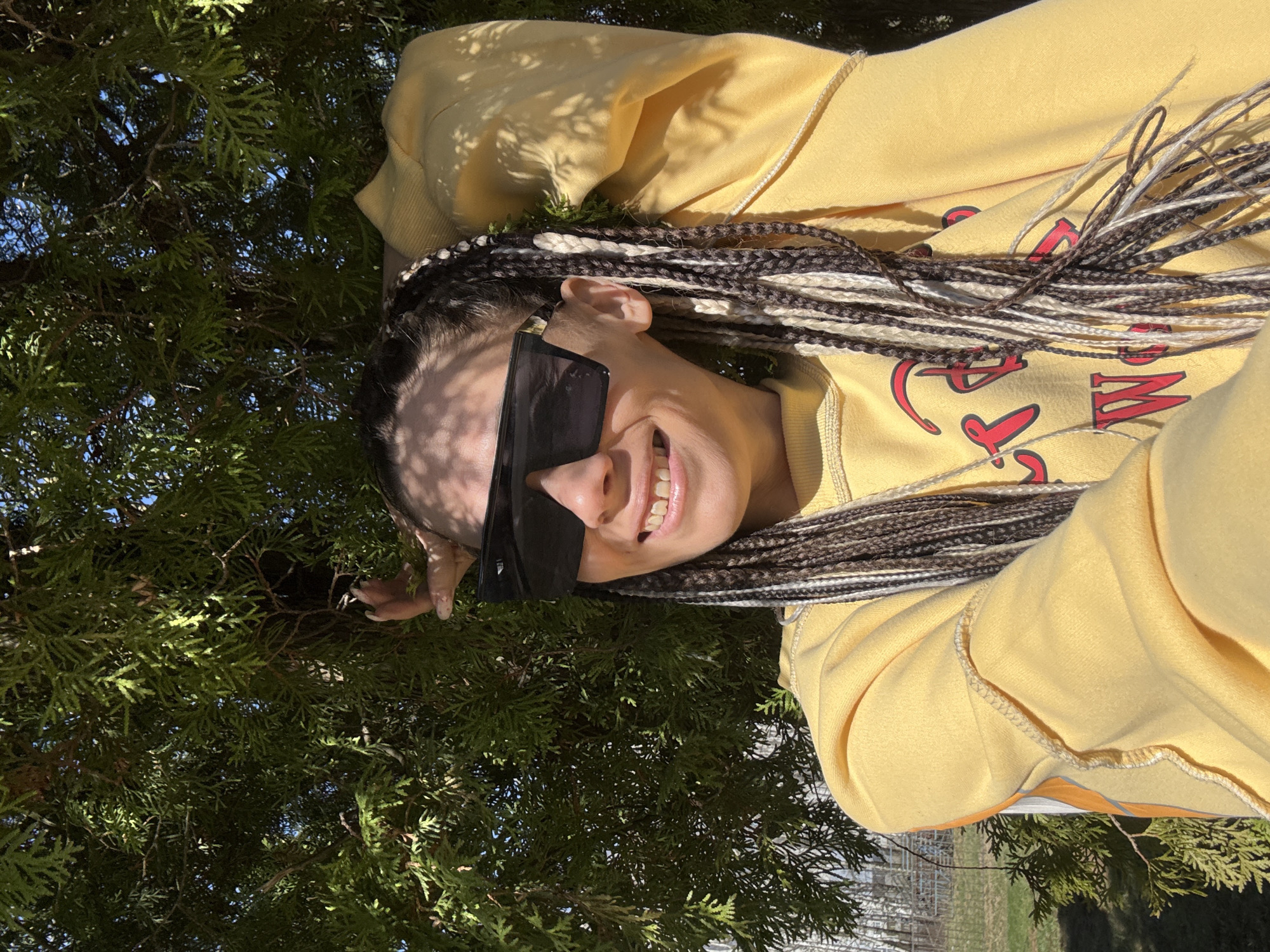 A young woman with dark and light braided hair is smiling at the camera. She is wearing sunglasses and a yellow sweater. The background is a green tree. The photo appears to have been taken outdoors in Rusanovo, Russia.