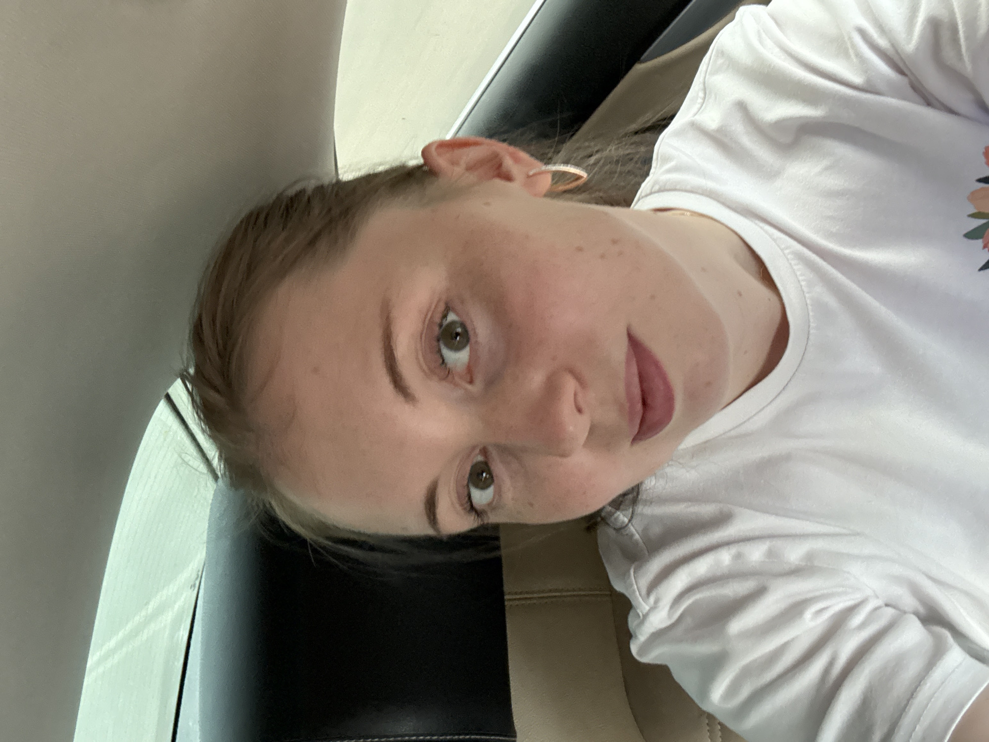 A close-up, selfie-style shot shows a young woman with light brown hair pulled back, looking directly at the camera. She has green eyes, light skin with freckles, and is wearing a white t-shirt. She has on a gold hoop earring in her right ear. The background shows the interior of a car, with a dark headrest and a lighter seat visible. The window behind her is slightly fogged, and a blurred outdoor scene is visible through it. The image appears to have been taken during the daytime, possibly while traveling. The location is Unknown, Unknown.