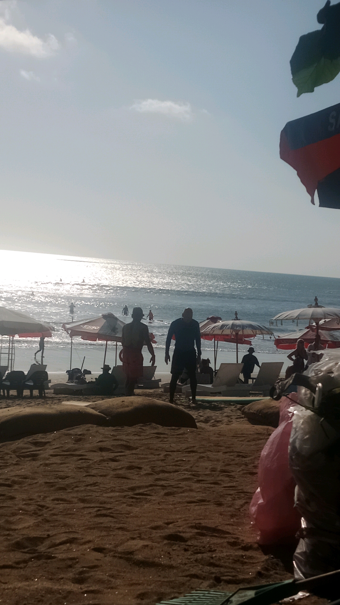 This is a medium shot, taken at eye level, of a busy beach scene in Kuta, Indonesia. The image is dominated by the ocean, which occupies the upper half of the frame, with the sun creating a shimmering reflection on the water. In the foreground, the sandy beach is littered with large, tan-colored beanbag-like objects. Two men, silhouetted against the bright background, stand near the center of the frame. The man on the left is wearing red shorts, and the man on the right is wearing dark shorts and a dark long-sleeved shirt. Behind them, several beach umbrellas, mostly red and white, are set up. People can be seen scattered across the beach, some sitting on lounge chairs, others walking or standing. In the water, numerous individuals are engaged in various activities, including surfing. The sky is light blue with a few wispy clouds. The overall impression is one of a lively, sunny day at the beach.