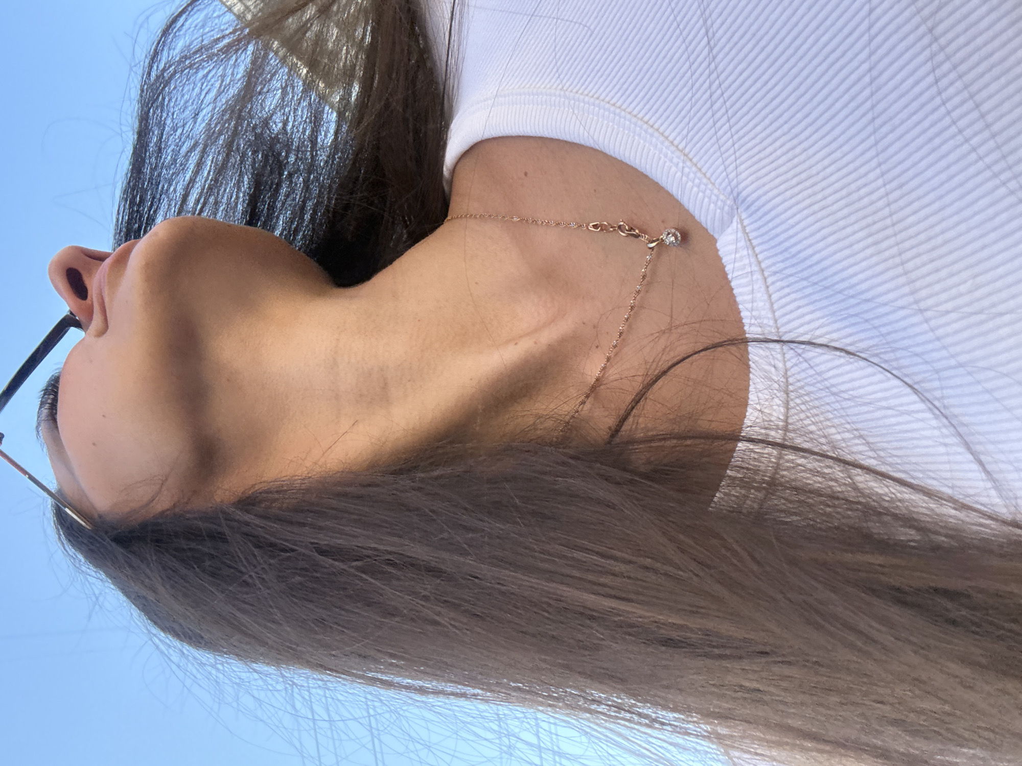 The image is a close-up, low-angle shot of a person's neck and chest, taken outdoors against a clear blue sky. The person is wearing a white ribbed tank top and a delicate rose gold necklace with a small diamond pendant. Their long, dark brown hair is blowing to the left, obscuring part of the frame. The person is looking upwards and to the right, with sunglasses visible at the top of the frame. There are no visible landmarks or architectural features to suggest a specific location within Kyiv, Ukraine.