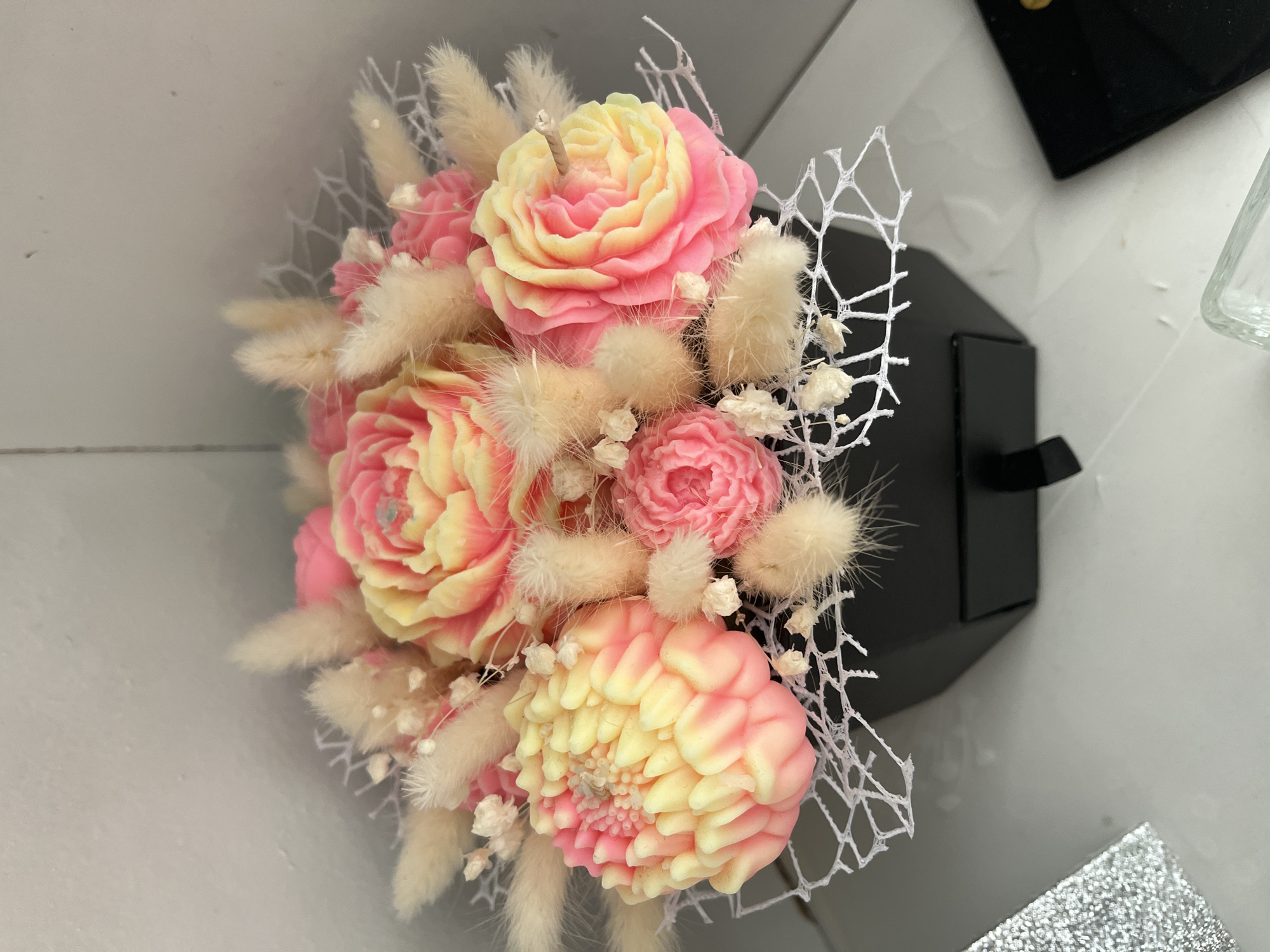 This media file displays a close-up, overhead view of a decorative bouquet of handcrafted soap flowers, presented in a black hexagonal box. The bouquet features several intricately carved soap flowers in shades of pink and yellow, resembling roses and possibly a dahlia. Interspersed among the flowers are fluffy, light-brown dried grasses and delicate white dried flowers, adding texture and a whimsical touch. A thin white netting wraps around a portion of the bouquet, enhancing its aesthetic appeal. A single white candle wick is visible at the center of the topmost flower, suggesting the bouquet may also function as a decorative candle. The background is a neutral, light-colored surface, possibly a table, with hints of other objects to the right, including a black rectangular item and a glass container, though these are out of focus. The lighting appears to be artificial and bright, creating soft shadows and highlighting the details of the soap flowers. The overall impression is one of delicate craftsmanship and aesthetic presentation, likely intended as a gift or decorative item. No people or specific location cues beyond the general setting are visible. There is no visible text in the media file.