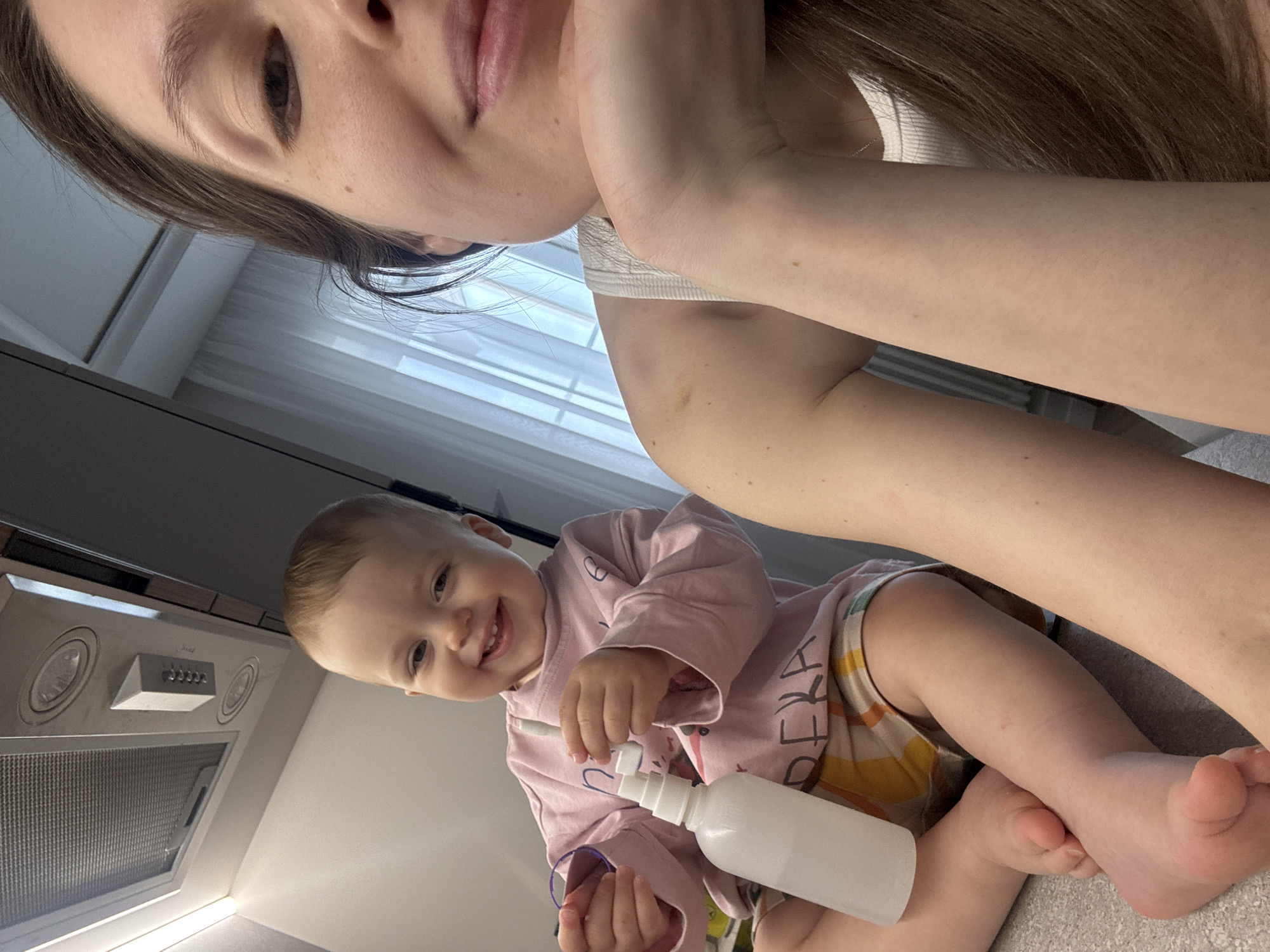 The image is a close-up, upward-angled selfie featuring a woman and a toddler in an indoor setting, likely a home kitchen in Moscow, Russia.
The upper right portion of the image shows the woman's face, with her left eye and part of her mouth visible. She has light brown hair and a calm expression. Her left arm is visible, resting on something out of frame, and she is wearing a white top.
Below and to the left, a smiling toddler, appearing to be about one to two years old, is looking directly at the camera. The toddler has light hair and is wearing a pink long-sleeved shirt and patterned shorts. In their right hand, the toddler holds a white spray bottle, and in their left hand, a purple elastic band. The toddler is sitting on a light-colored, textured surface.
In the background, a modern kitchen range hood with metallic finishes, round lights, and a control panel is visible, suggesting a kitchen environment. A window with horizontal blinds provides natural light, indicating it is daytime. On the toddler's shorts, visible Cyrillic letters form "ЕКЛА". The overall mood is cheerful, primarily conveyed by the toddler's wide smile and engaged expression during what appears to be a playful interaction.