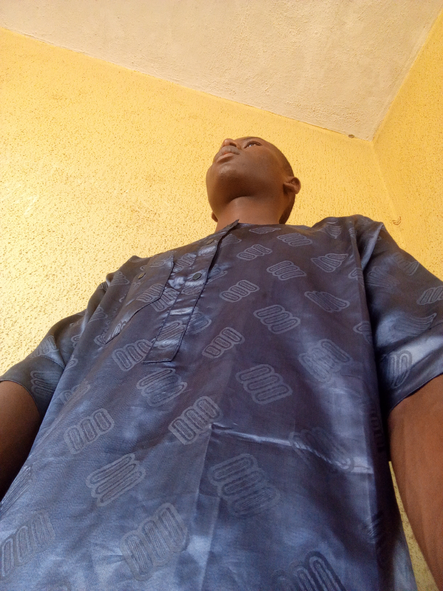 A low-angle shot depicts a dark-skinned man wearing a patterned blue shirt. He is gazing upwards. The background consists of a textured, yellow-painted wall. The location is unspecified in the image, but inferred based on the prompt.