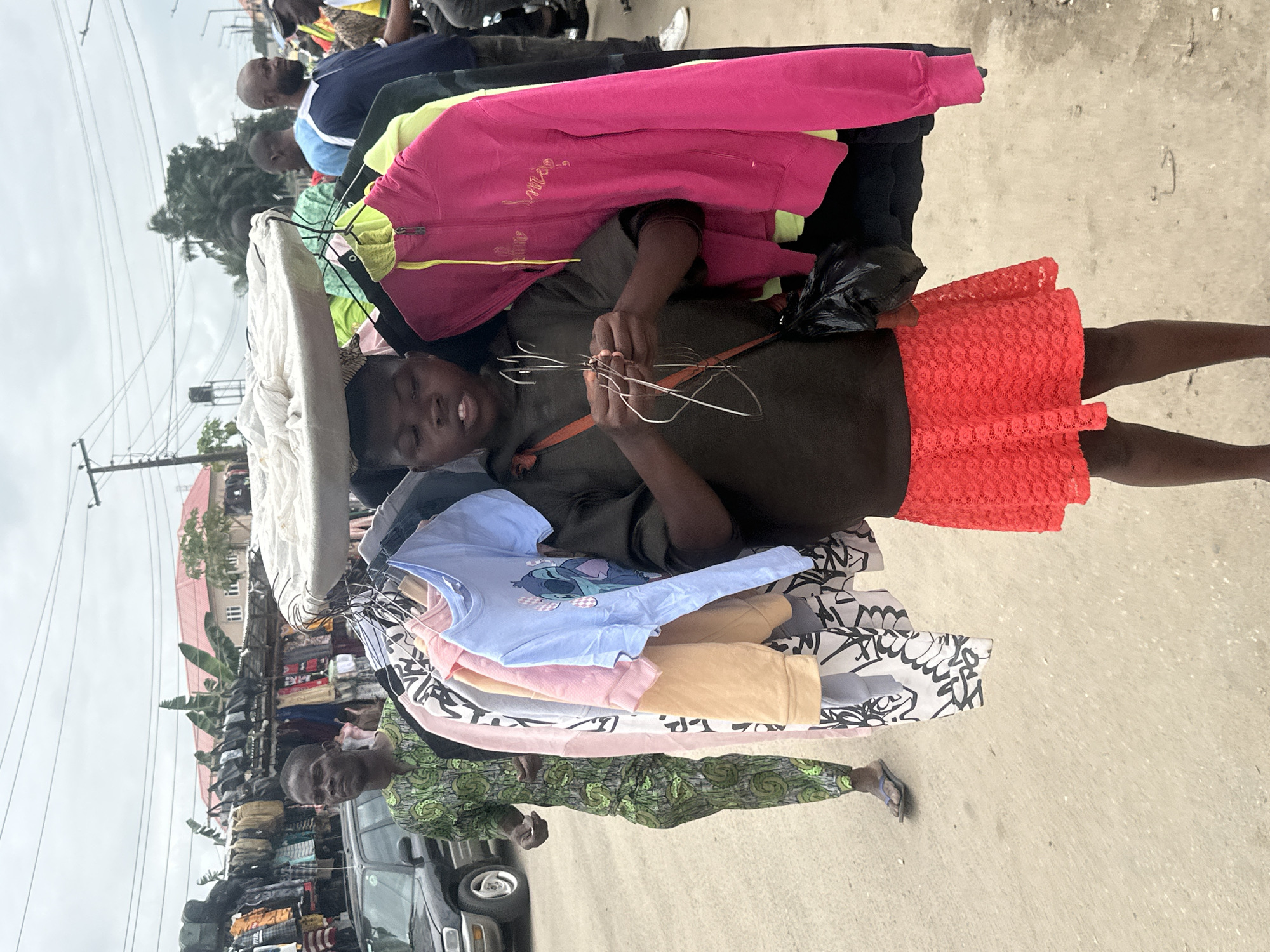 A young person carries a display of clothing on their head in a busy street scene in Badagry, Nigeria. The individual is wearing a dark long-sleeved shirt and a bright red lace skirt. They are holding a bundle of wire hangers in their hands, suggesting they are a street vendor. The clothing displayed on hangers includes a light blue t-shirt with a cartoon character, a pink hoodie, and a bright pink zip-up jacket. Behind the young person, a man in a green patterned dashiki walks away from the camera, and a black car is parked on the left. The environment appears to be a roadside market, with stalls and other people visible in the background. The ground is paved with asphalt or concrete, and the sky is overcast. Power lines and utility poles are visible overhead.