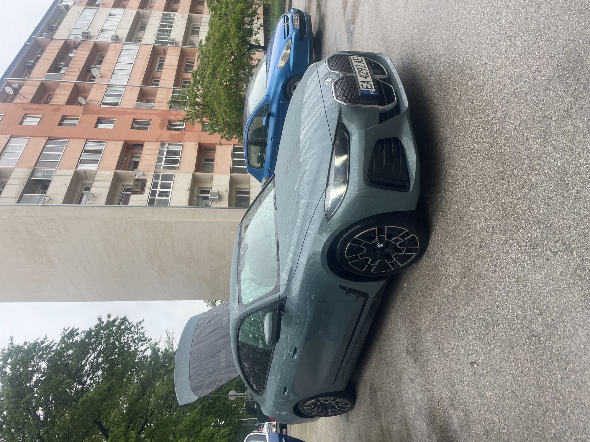 The image shows two cars parked on an asphalt surface in Gabrovo, Bulgaria. A gray BMW with license plate "EA 4292 AE" is parked in the foreground. Behind it, a blue Peugeot 307 with license plate "EB 0351 BB" is also parked. The hood of the BMW is open. In the background is a multi-story apartment building with visible air conditioning units. Green trees are also visible. The scene appears wet, possibly after rain.