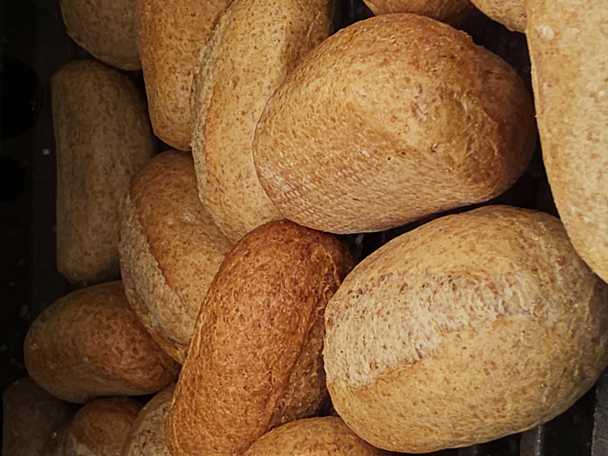 The image displays a close-up, top-down view of numerous golden-brown bread rolls. These rolls are oblong or oval-shaped and appear to be whole wheat or multigrain, characterized by their textured, slightly coarse surface and varying shades of brown. Many of the rolls exhibit subtle scoring lines on their crusts. They are densely packed and overlapping, suggesting a large quantity.
The background is dark and indistinct, possibly a baking sheet, oven rack, or a shadowy container, creating a strong contrast that highlights the bread. The lighting emphasizes the texture and color of the rolls, making them appear freshly baked. There are no people visible, and no specific activity beyond the apparent display or storage of the bread. The scene suggests an indoor setting, likely a bakery or a commercial kitchen, where the rolls are either cooling or awaiting sale. No text is visible in the image.