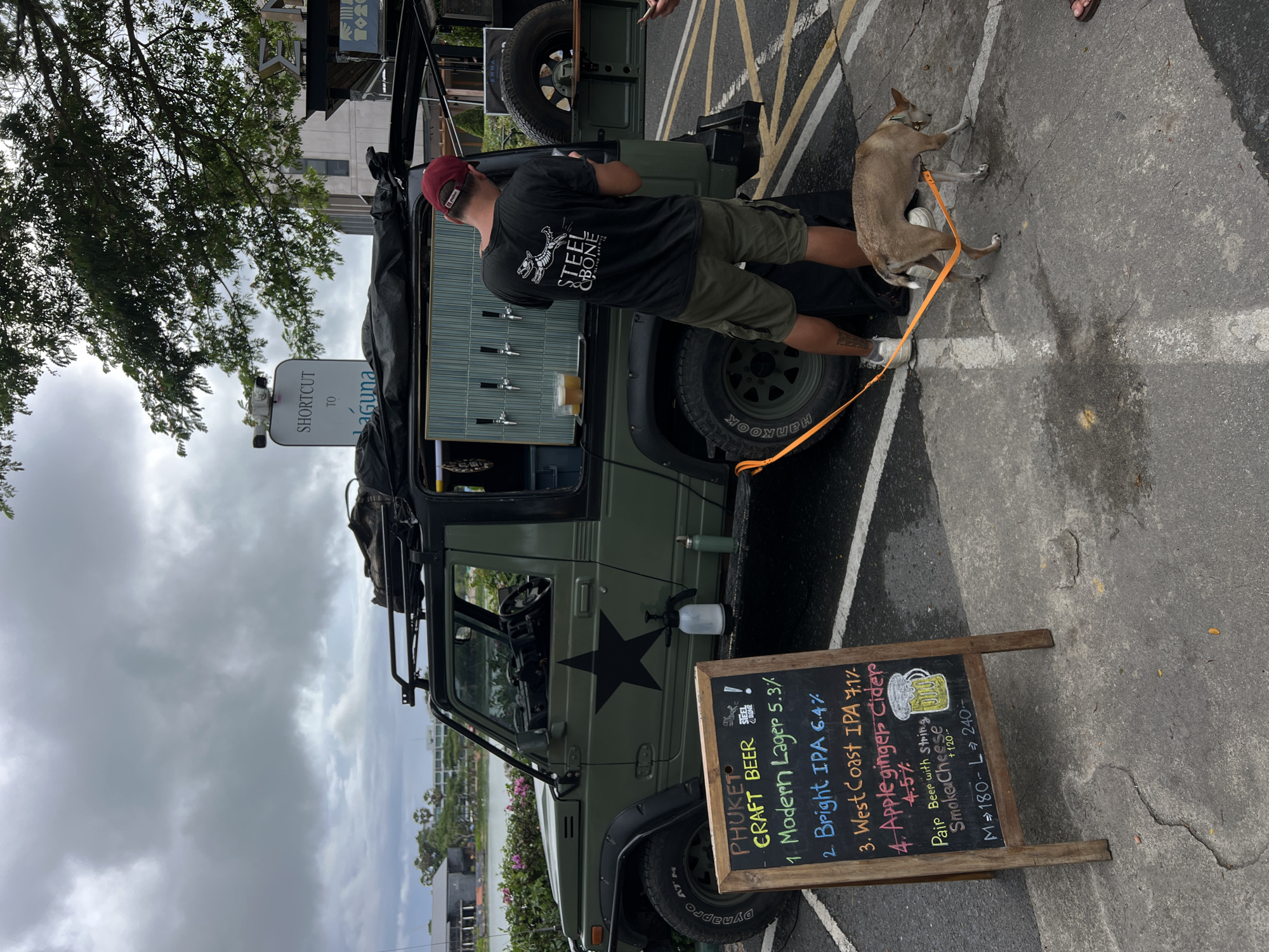 In an outdoor setting with an overcast sky, a man wearing a maroon baseball cap and a black t-shirt with "STEEL & BONE ISLAND BREW" printed on it, stands beside a modified olive-green Suzuki Samurai. The vehicle has been outfitted as a mobile bar with taps dispensing beverages. A small tan dog on an orange leash is being held by the man, who is looking down at the dog. A chalkboard sign displaying "PHUKET CRAFT BEER" with a list of beers and prices stands in front of the vehicle. The background shows a body of water, buildings, and trees. The ground is paved with asphalt and marked with white lines.