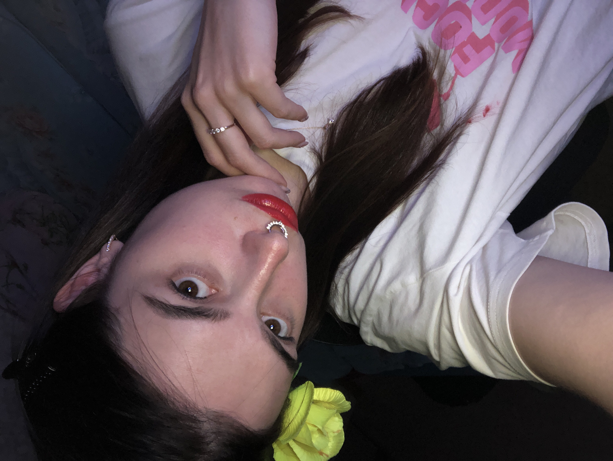 A young woman is pictured from a high angle, lying on her back. She is wearing a white t-shirt with pink writing. She has brown hair and is wearing red lipstick and a nose ring. A yellow flower is tucked near her left ear. She is wearing a diamond ring on her ring finger and a necklace. The background is dark and out of focus, but a blue patterned couch is partially visible. There is no evidence to suggest the location is Noviy, Russia.