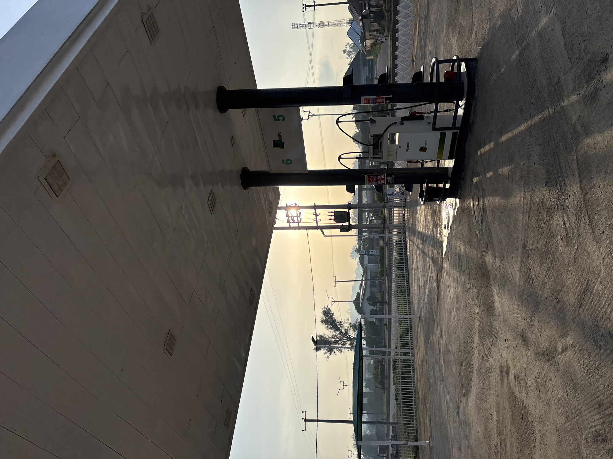 The image depicts a gas station with a prominent canopy. The canopy's underside is textured with a grid pattern and features embedded square lights or vents. Two thick, dark, cylindrical pillars support the canopy. Beneath the canopy, a white petrol pump with "NNPC" branding and a "PETROL" label is visible, alongside two dispensing nozzles. The pump is situated on a black platform with metal railings. The ground around the pump is a concrete or asphalt surface with visible tire marks and some wet patches reflecting the sunlight.
In the background, through the canopy's supports and the open space, a hazy, sunlit sky is visible. The sun is low on the horizon, casting long shadows across the ground. Several utility poles with wires are scattered throughout the scene. A white picket fence runs along the edge of the gas station's forecourt. Beyond the fence, there are buildings and trees, suggesting a semi-urban or rural environment. The overall impression is of an outdoor scene at either sunrise or sunset, with a focus on the functional elements of a gas station. The location is identified as Bukuru, Nigeria.