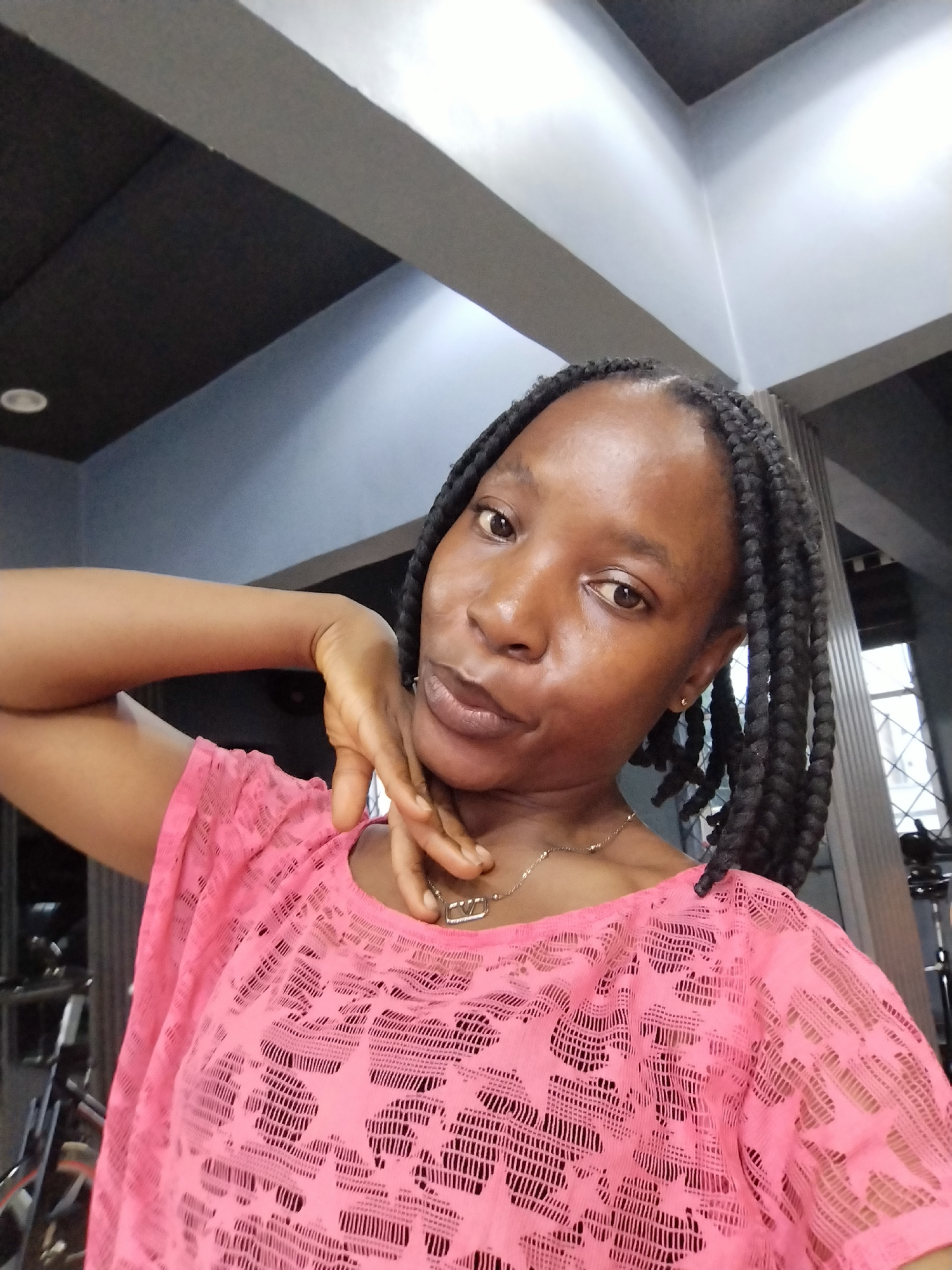 In Iguosa, Nigeria, a young Black woman with braided hair and a pink, lace-like top poses for a selfie. Her left arm is bent at the elbow, and her left hand is positioned on her jawline, with her fingers gently touching her cheek. She wears a delicate gold necklace with a small, square pendant. The background suggests an indoor setting, possibly a gym or studio, with muted gray and black architectural elements, including ceiling beams and what appear to be gym equipment in the background. Sunlight streams in from a window on the right.
