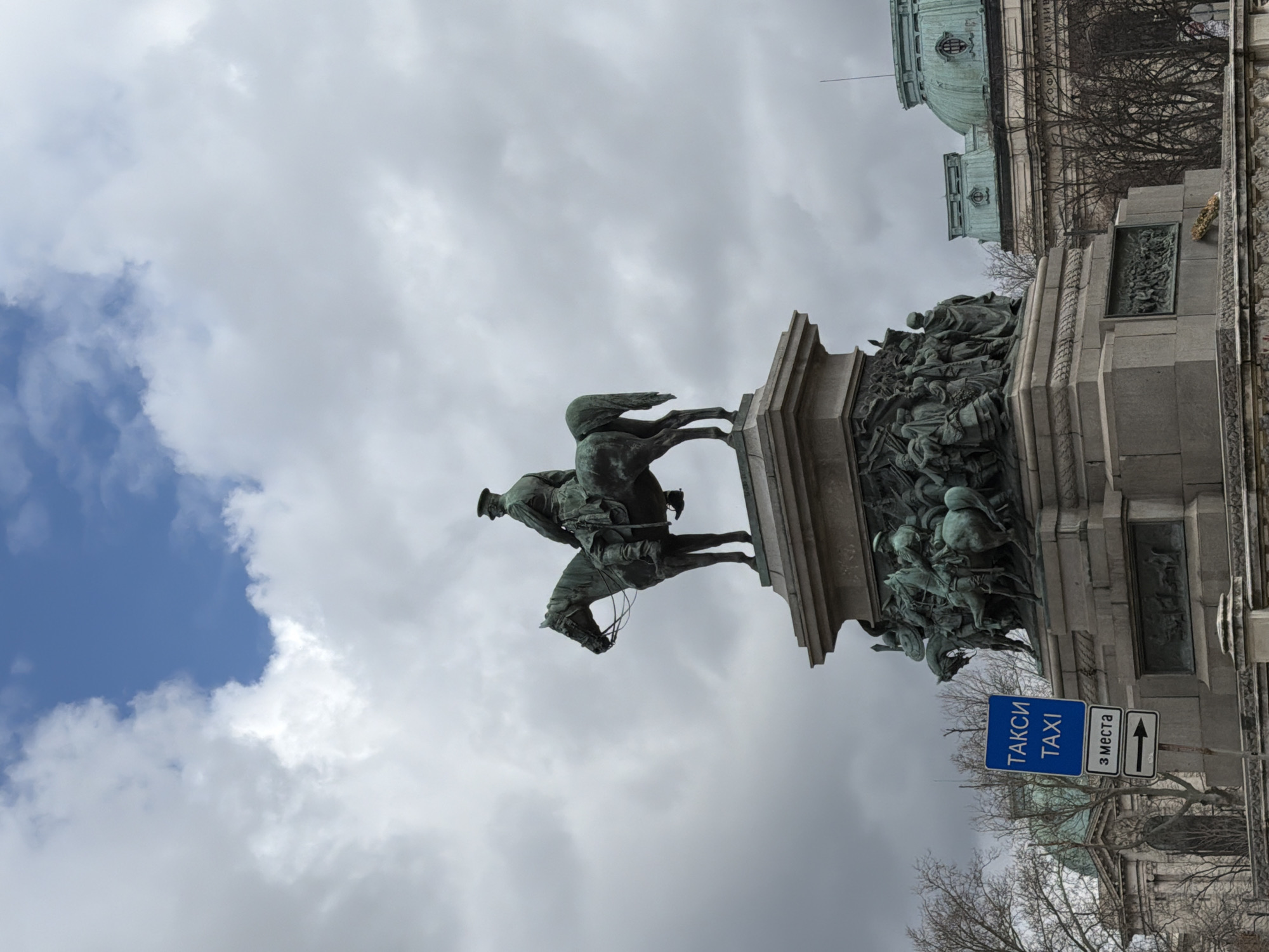The image shows a statue in a city, presumably Unknown, Unknown. The statue depicts a man on horseback atop a highly decorated base. The surrounding area includes a blue sign with white Cyrillic and Latin lettering, indicating a taxi stand with three spaces. Bare trees are visible in the background along with what appears to be a building with a green dome. The sky is overcast with patches of blue showing through the clouds.