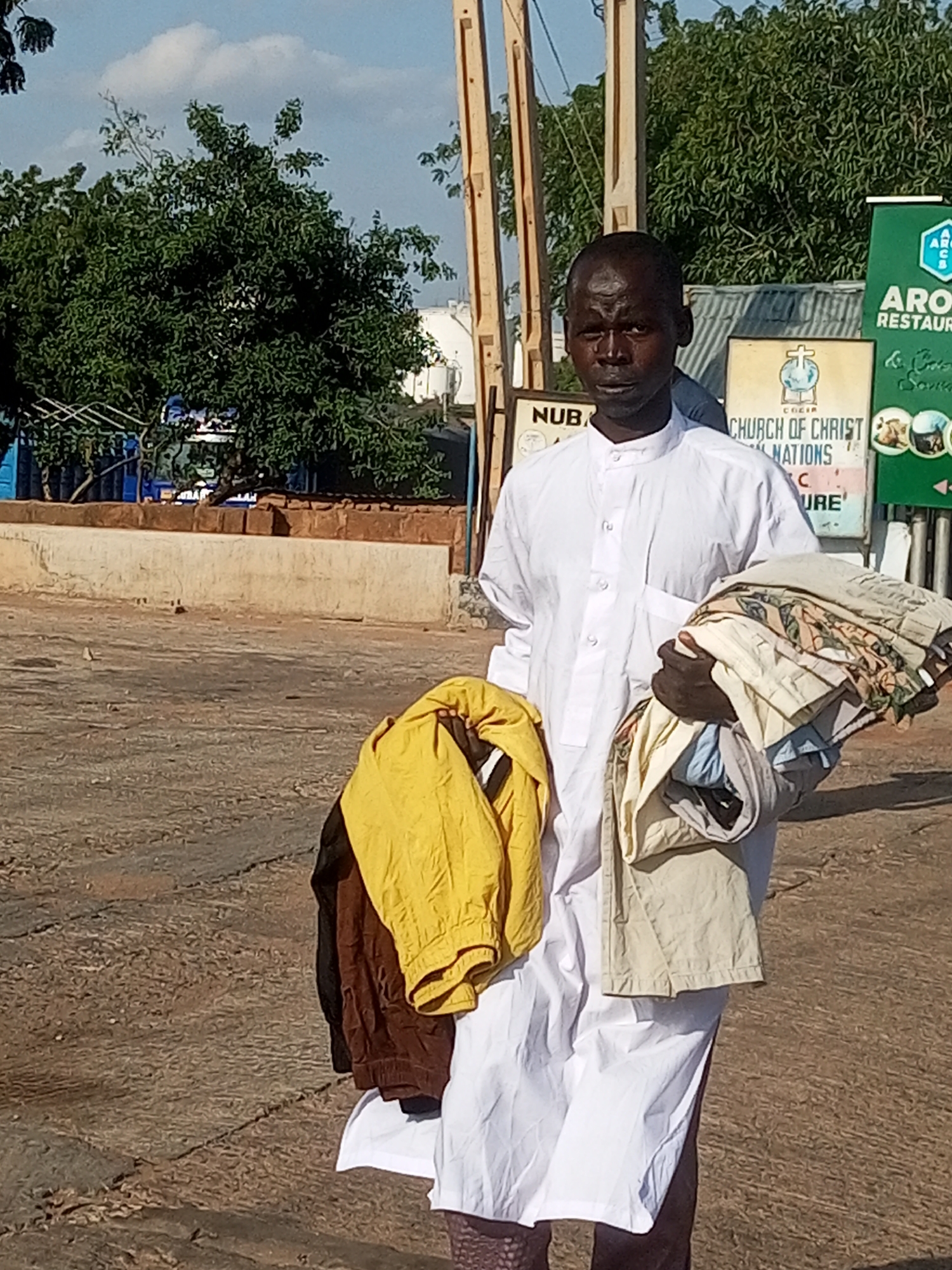 A man wearing a white robe stands outdoors holding folded clothes. He is carrying a bundle of fabric in his right arm and a yellow garment over his left arm. Behind him, signs are visible, including one that reads "NUBA" and another for "CHURCH OF CHRIST NATIONS". A green sign with the text "ARO RESTAUR" is also present. Trees and a blue structure are in the background. The setting appears to be a street or public area with a paved surface. The lighting suggests it is daytime, likely in the morning or afternoon. The man's expression is neutral.